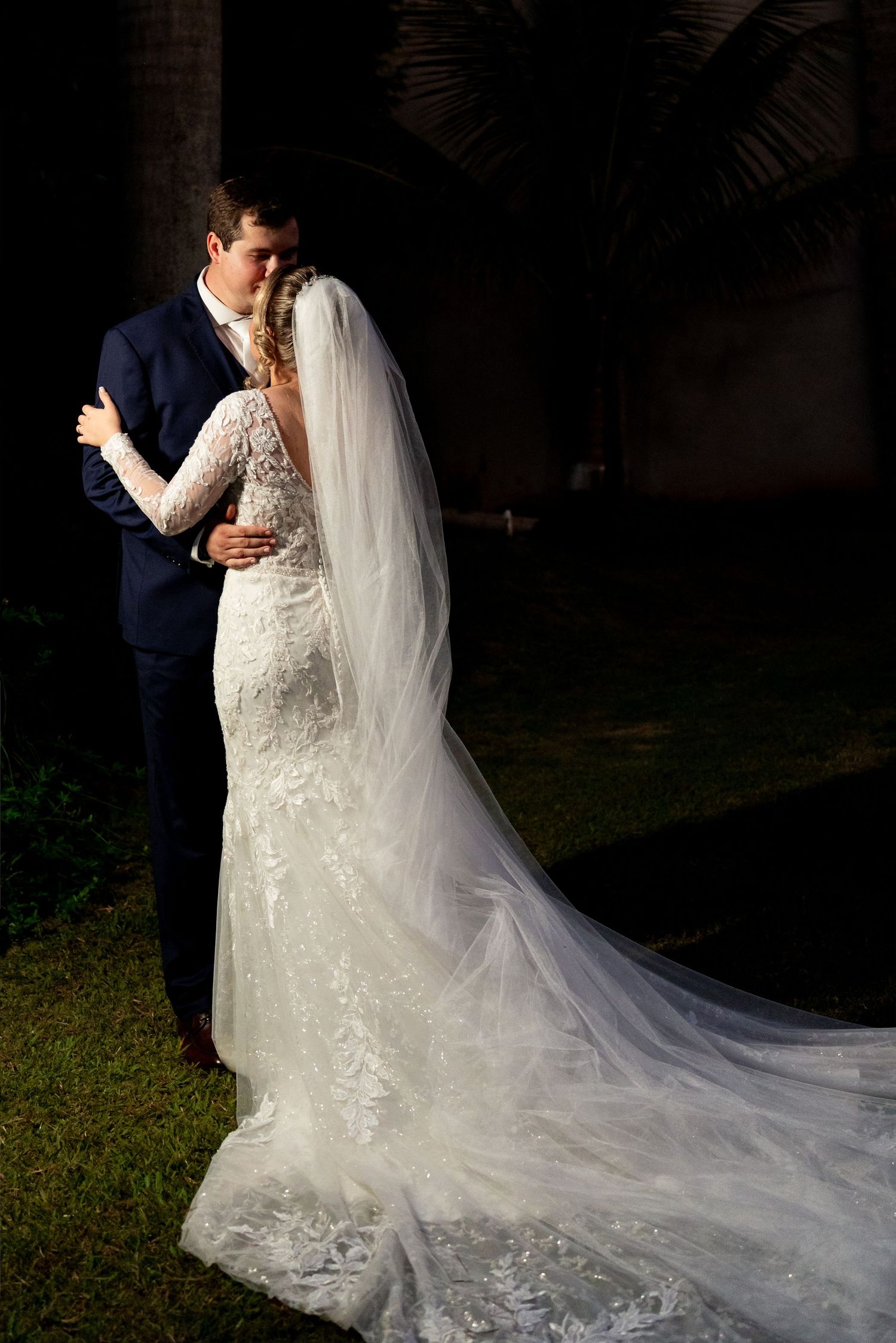 Bride in lace mermaid gown with veil and groom in navy suit share romantic kiss outdoors at wedding.