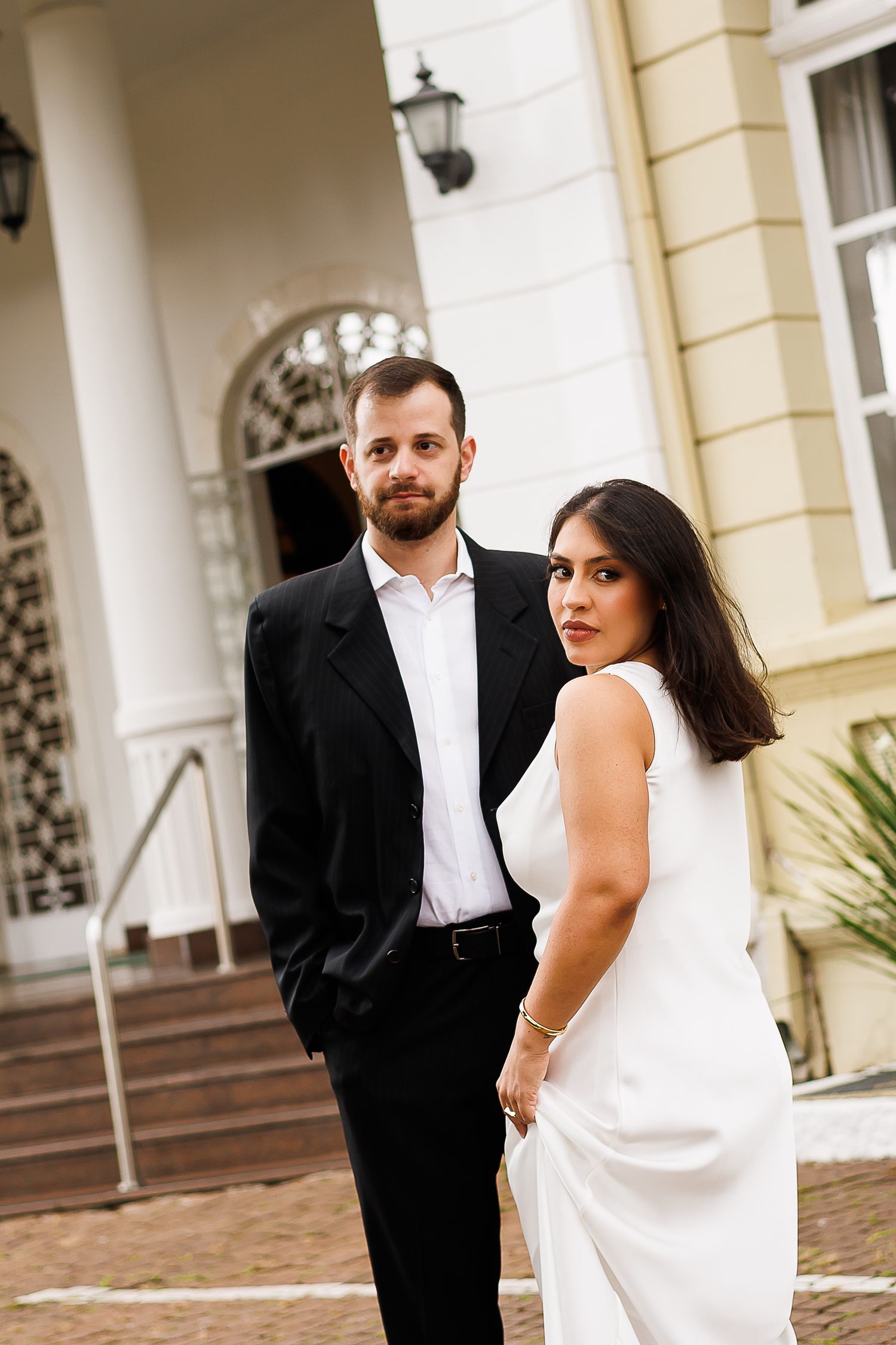 Elegant couple posing outdoors, man in black suit and woman in white dress, standing before a classic building entrance.