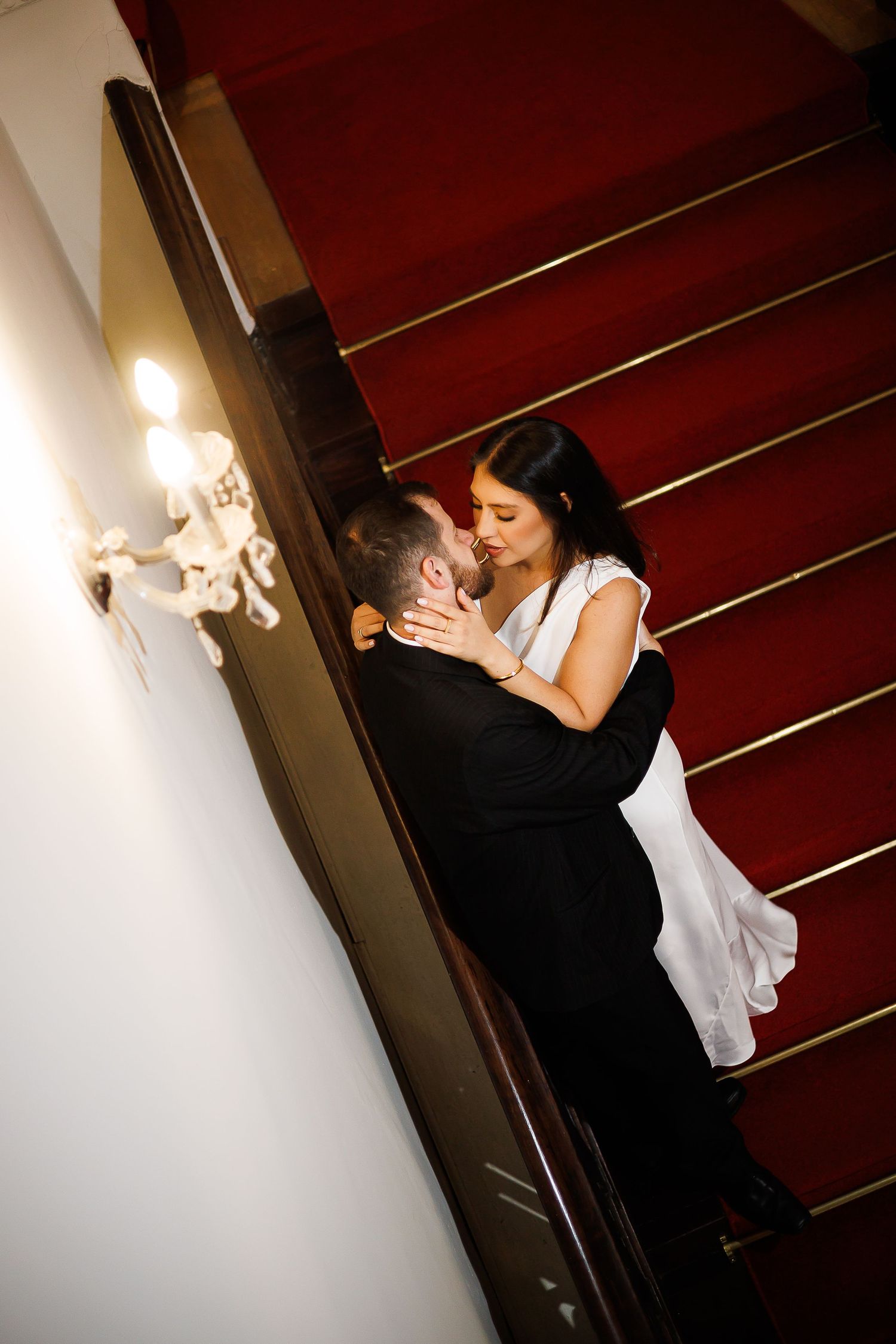 Bride and groom share a romantic moment on a grand staircase with red carpet and elegant wall sconce lighting.