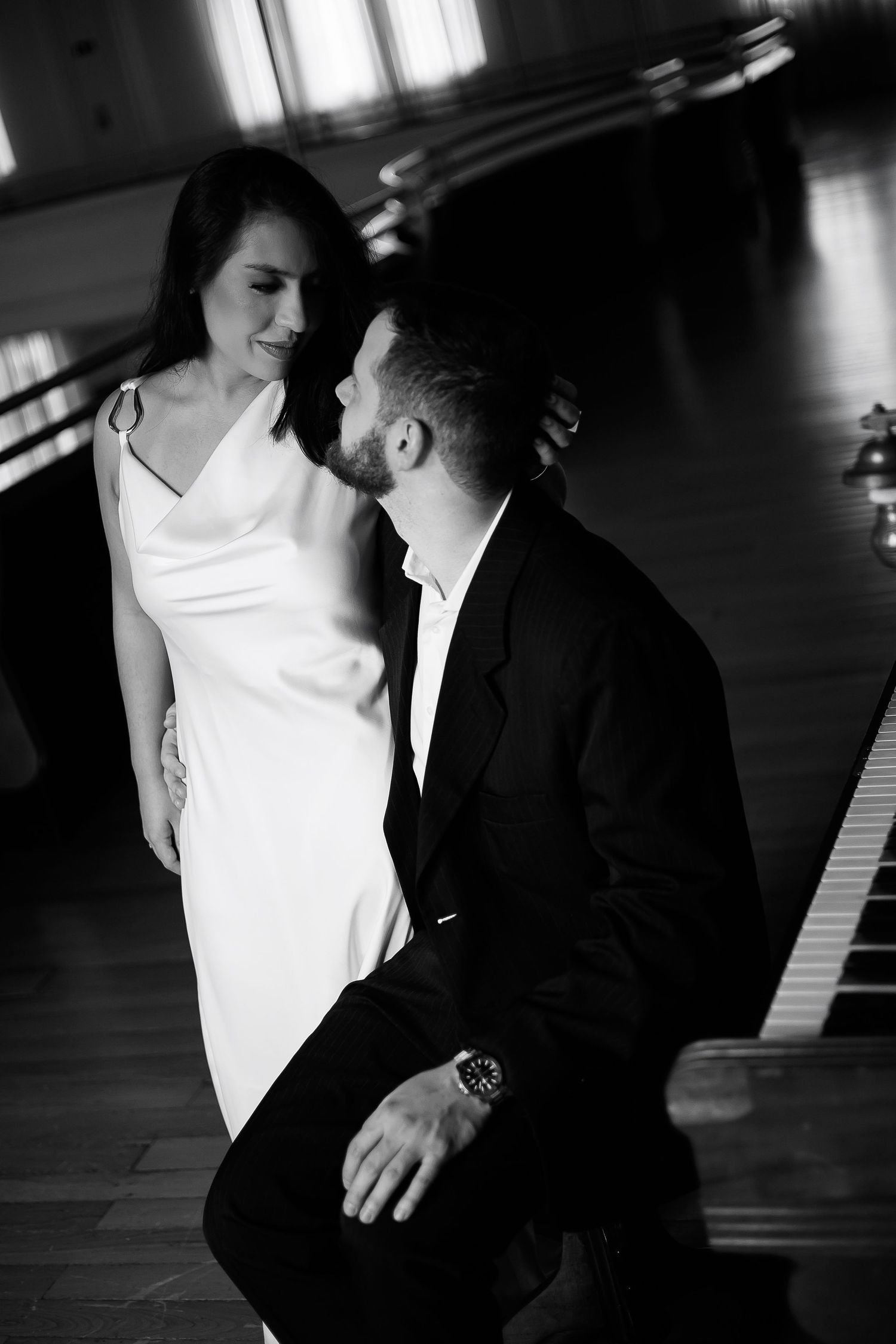 Black and white photo of a romantic couple sharing a tender moment near a grand piano in an elegant hall.