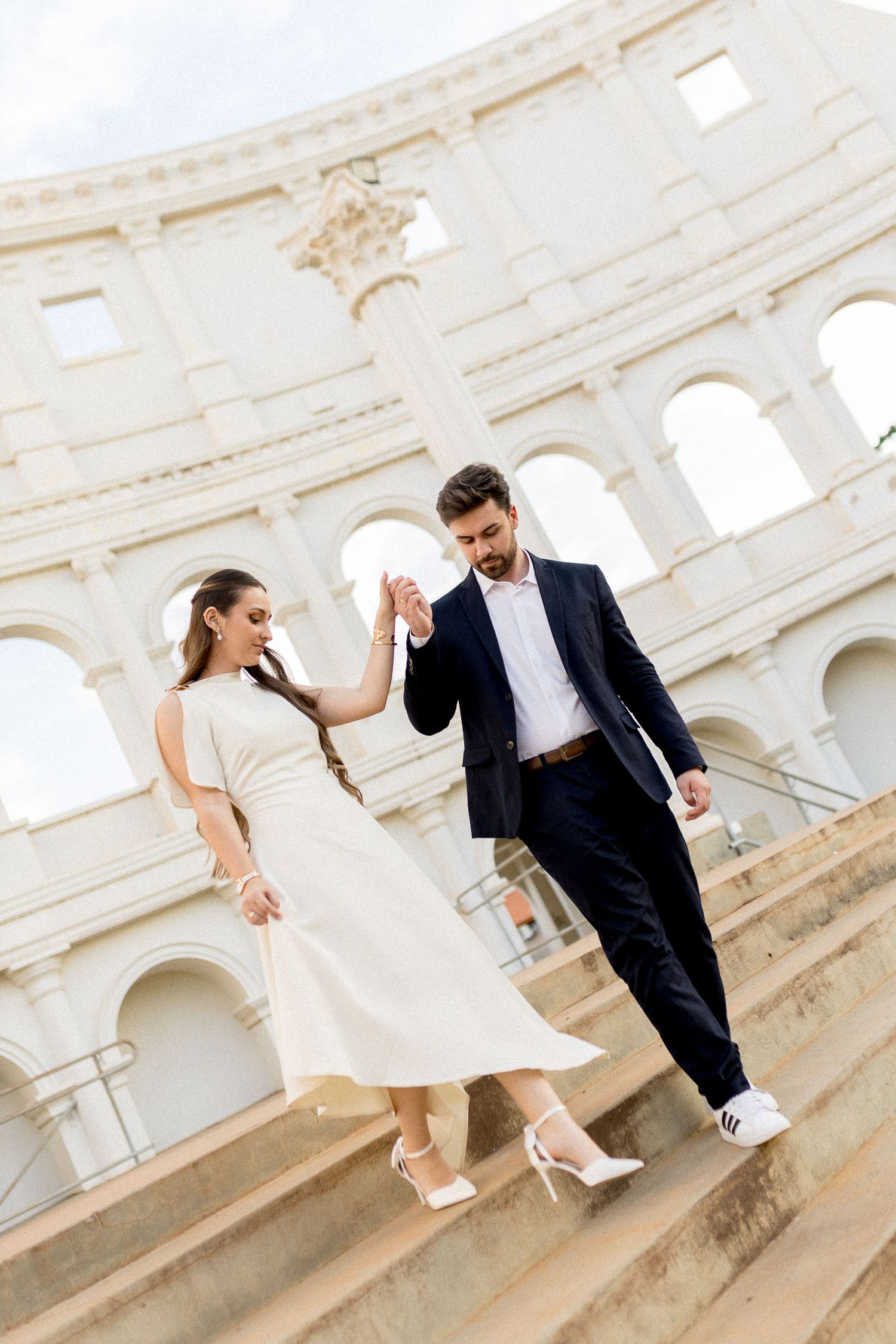 Elegant couple holding hands on steps before the Colosseum in Rome, woman in white dress, man in navy suit.