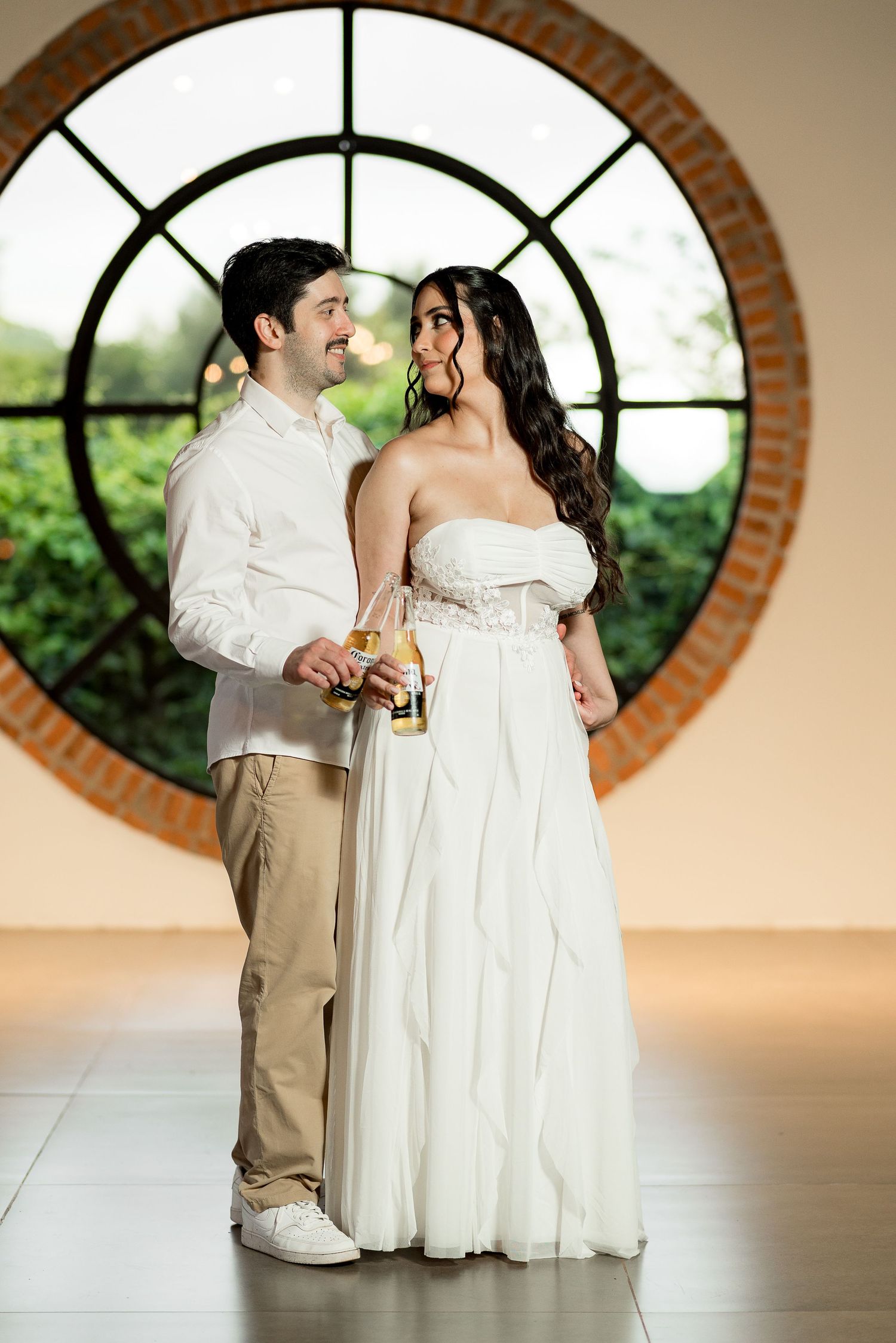 A couple in wedding attire smiles at each other holding drinks before a circular brick window.