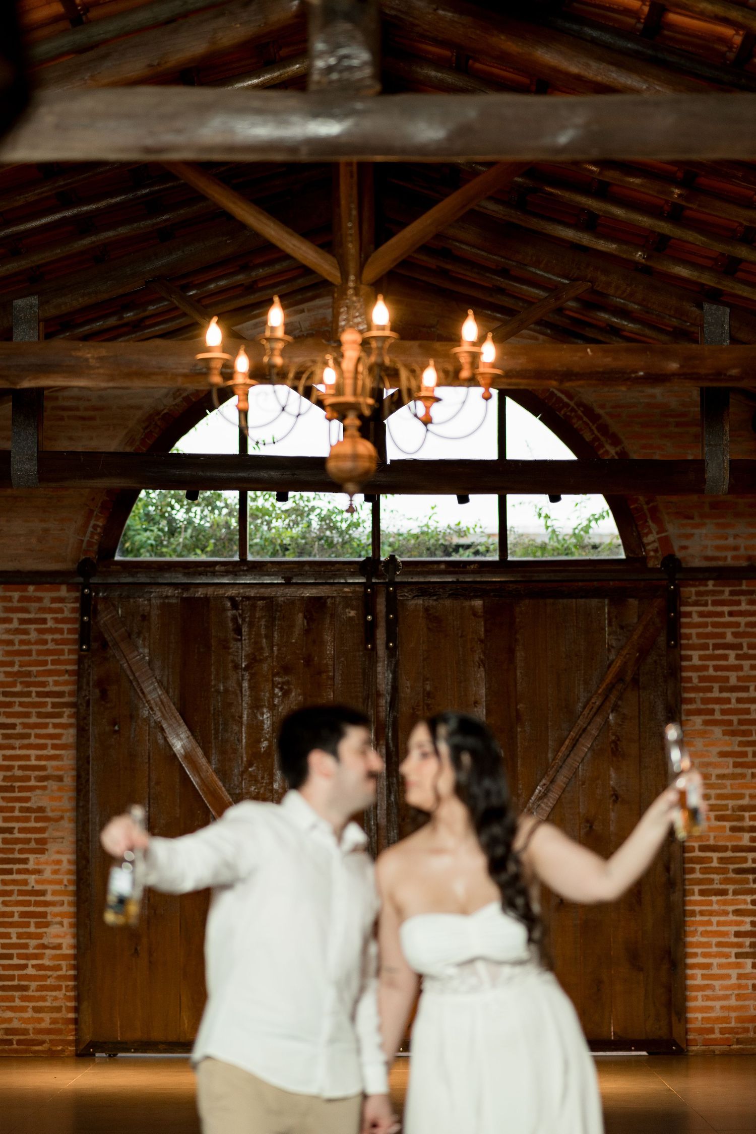 Couple in wedding attire dancing in a rustic barn venue with chandelier and wooden doors.