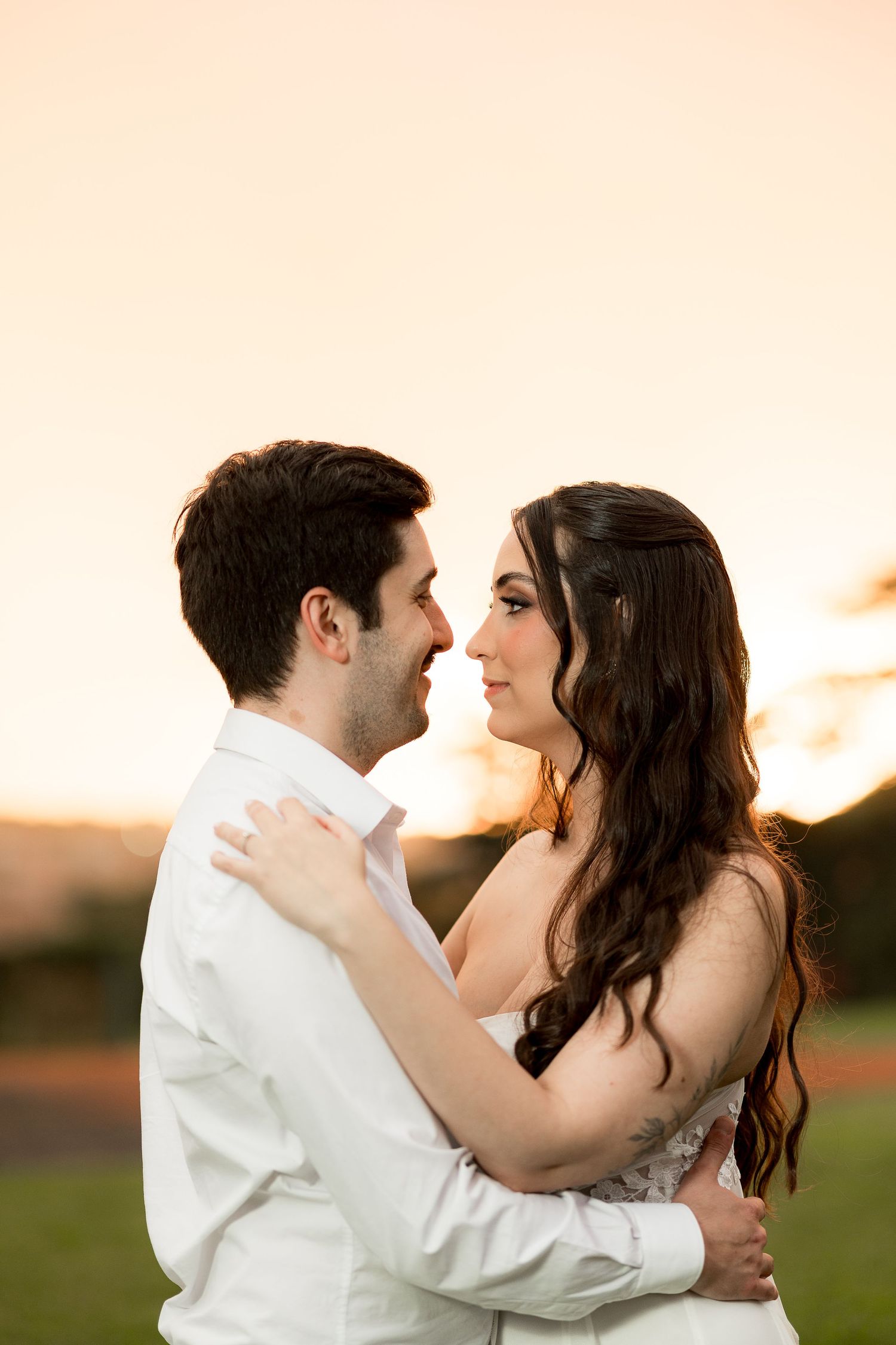 Couple embracing at sunset, bride in lace gown and groom in white shirt, sharing a romantic outdoor moment.