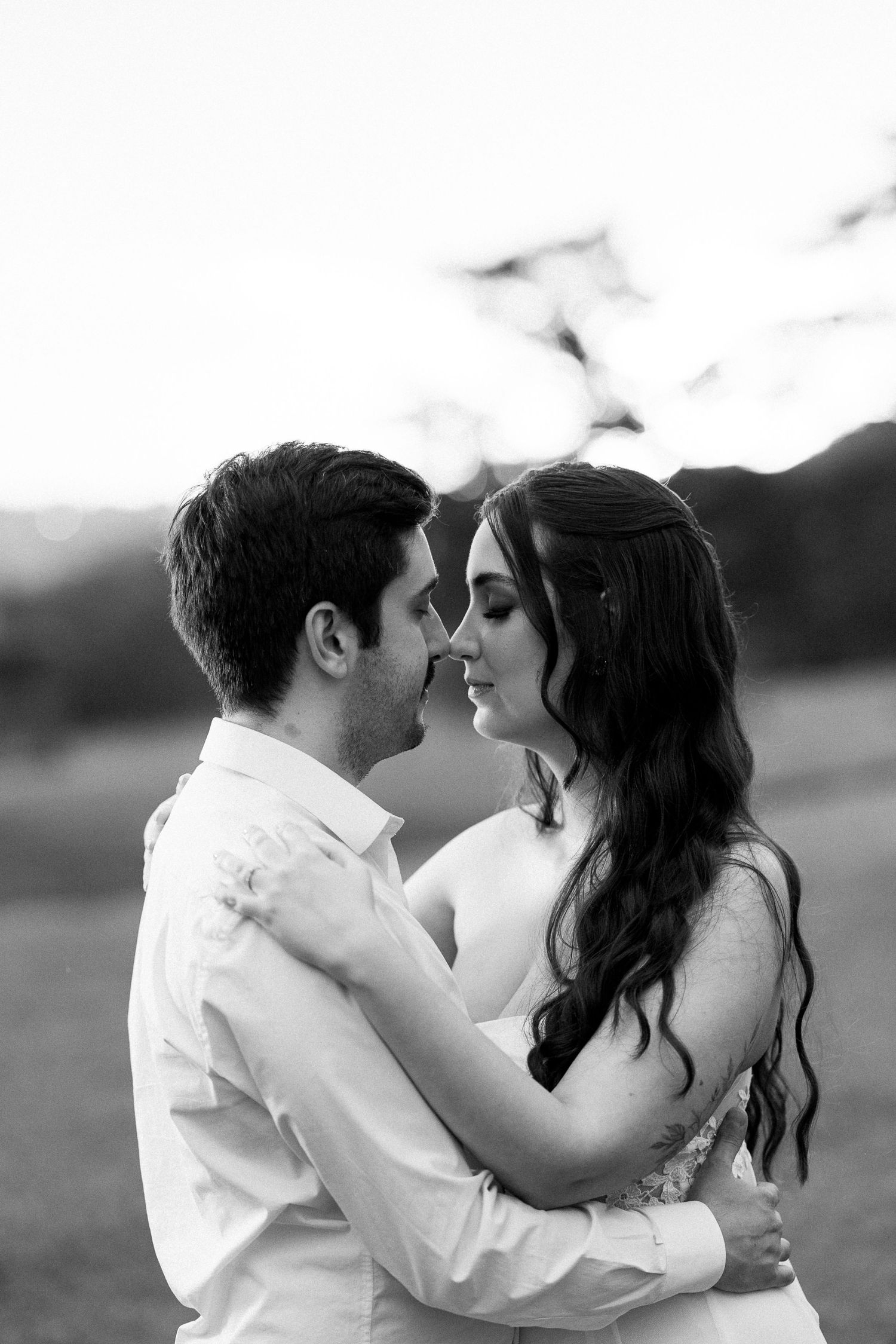 Black and white photo of a romantic couple embracing and leaning in for a kiss outdoors at sunset.