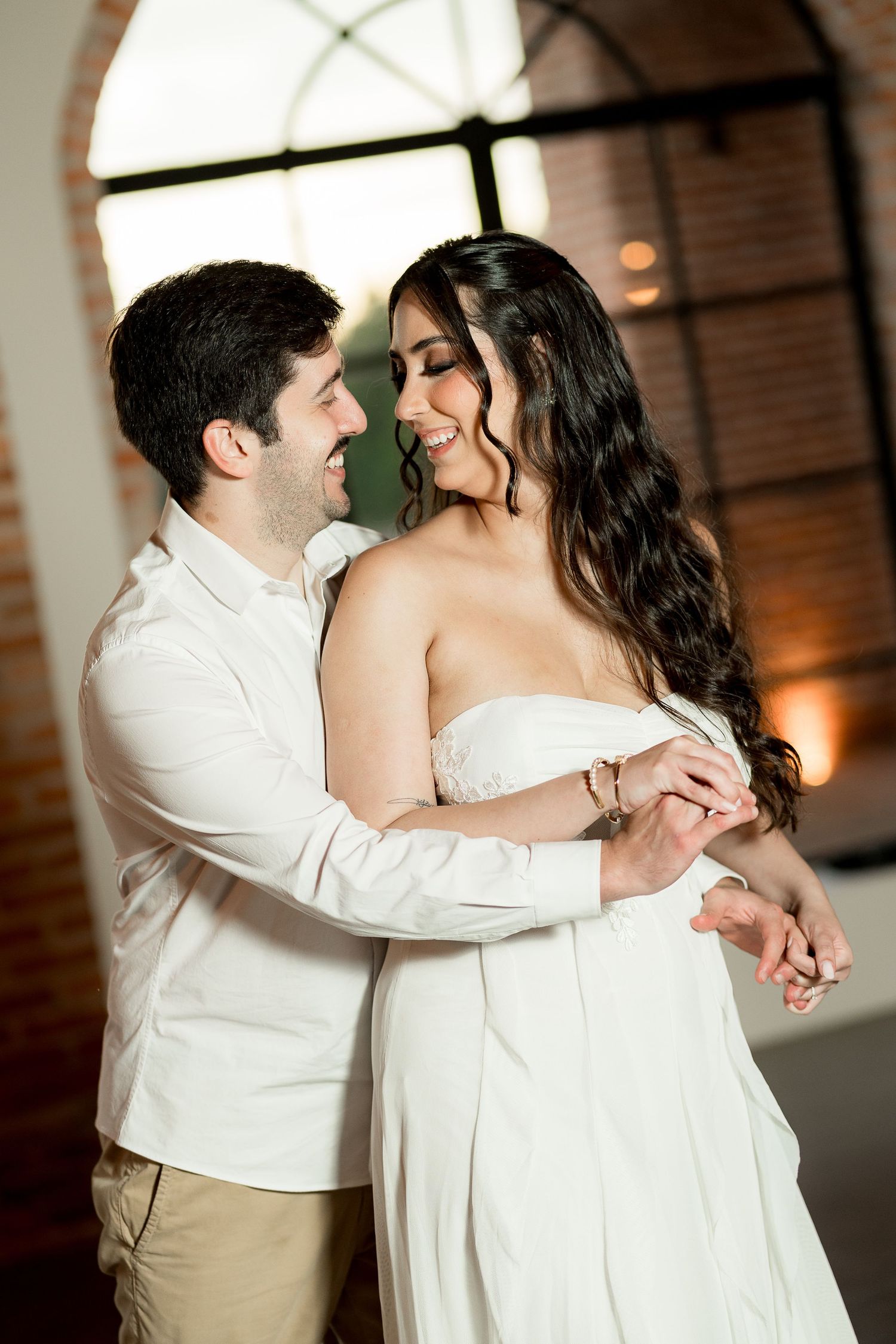 A couple shares a romantic first dance, the bride in a white off-shoulder gown and groom in white shirt, smiling joyfully.