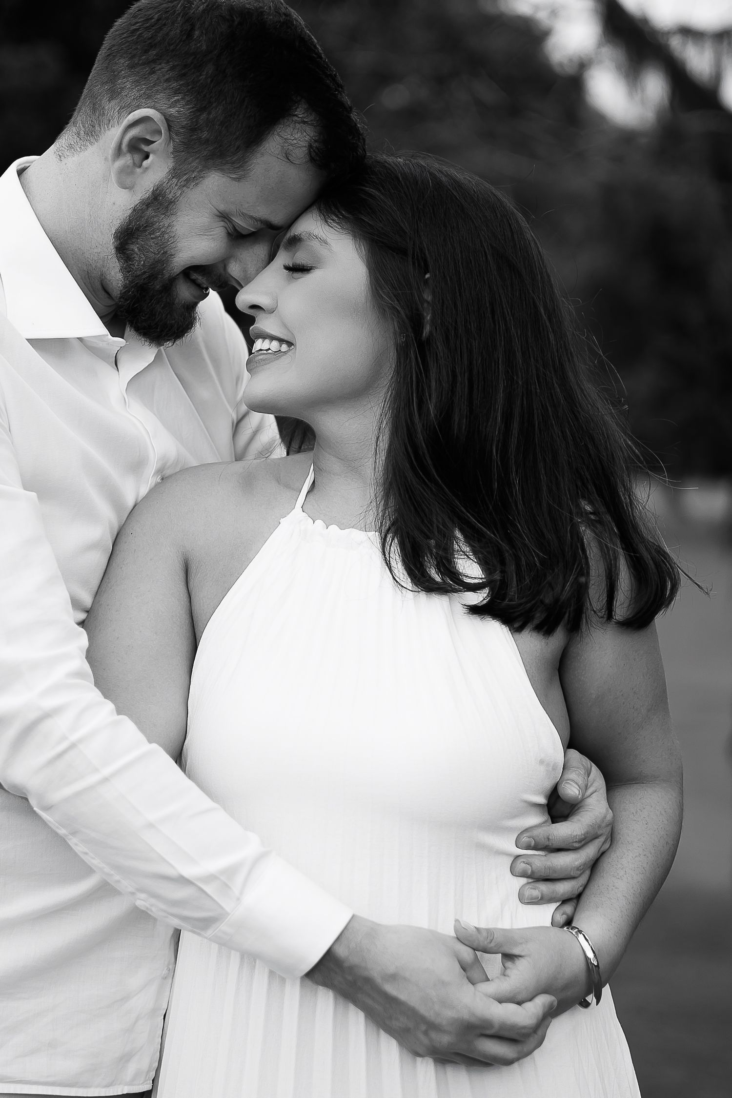 Black and white photo of a romantic couple embracing outdoors, woman in white dress smiling as man holds her close.