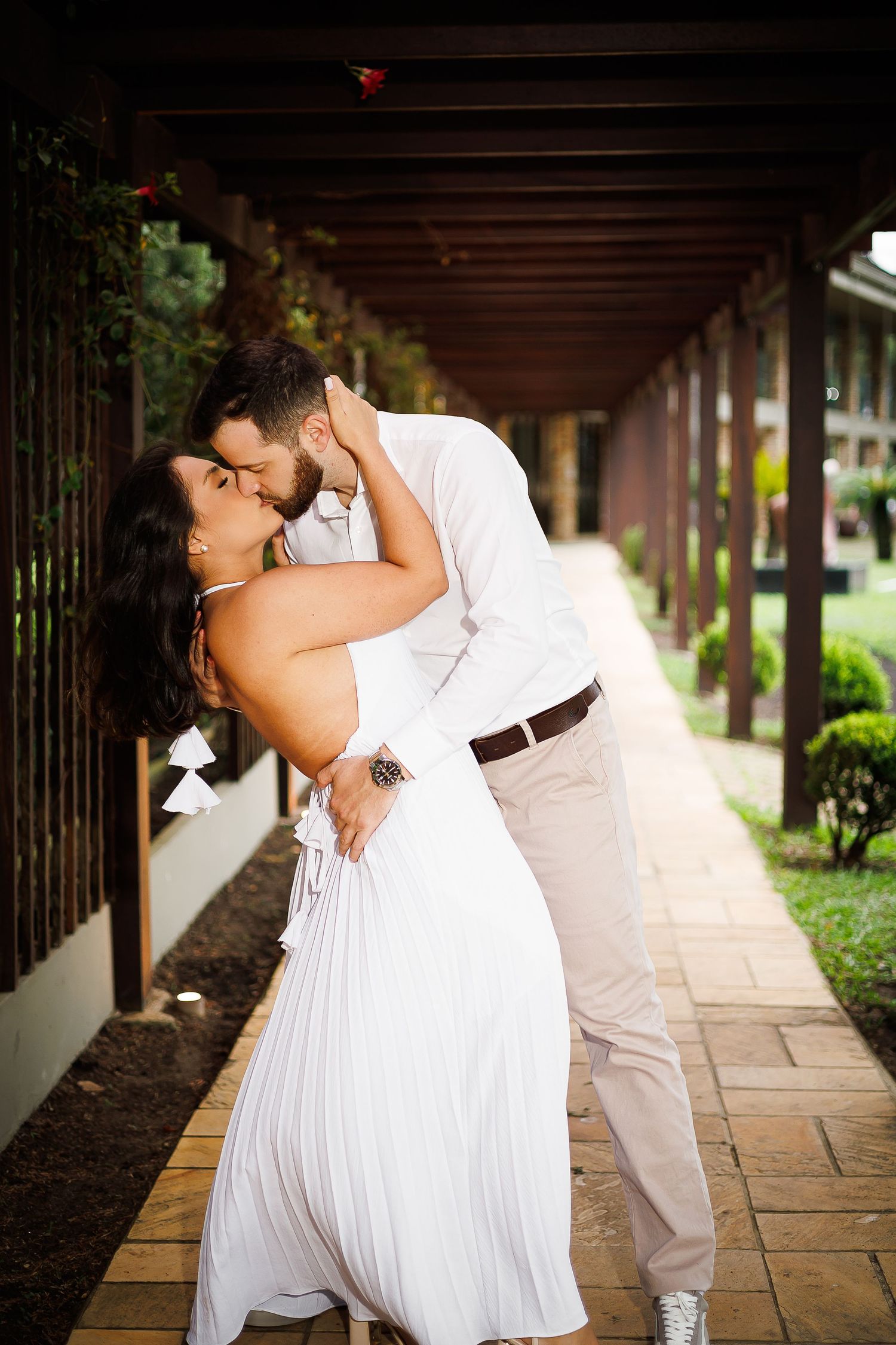 Couple sharing a romantic dip kiss under a wooden pergola, dressed in white outfits, surrounded by lush greenery.