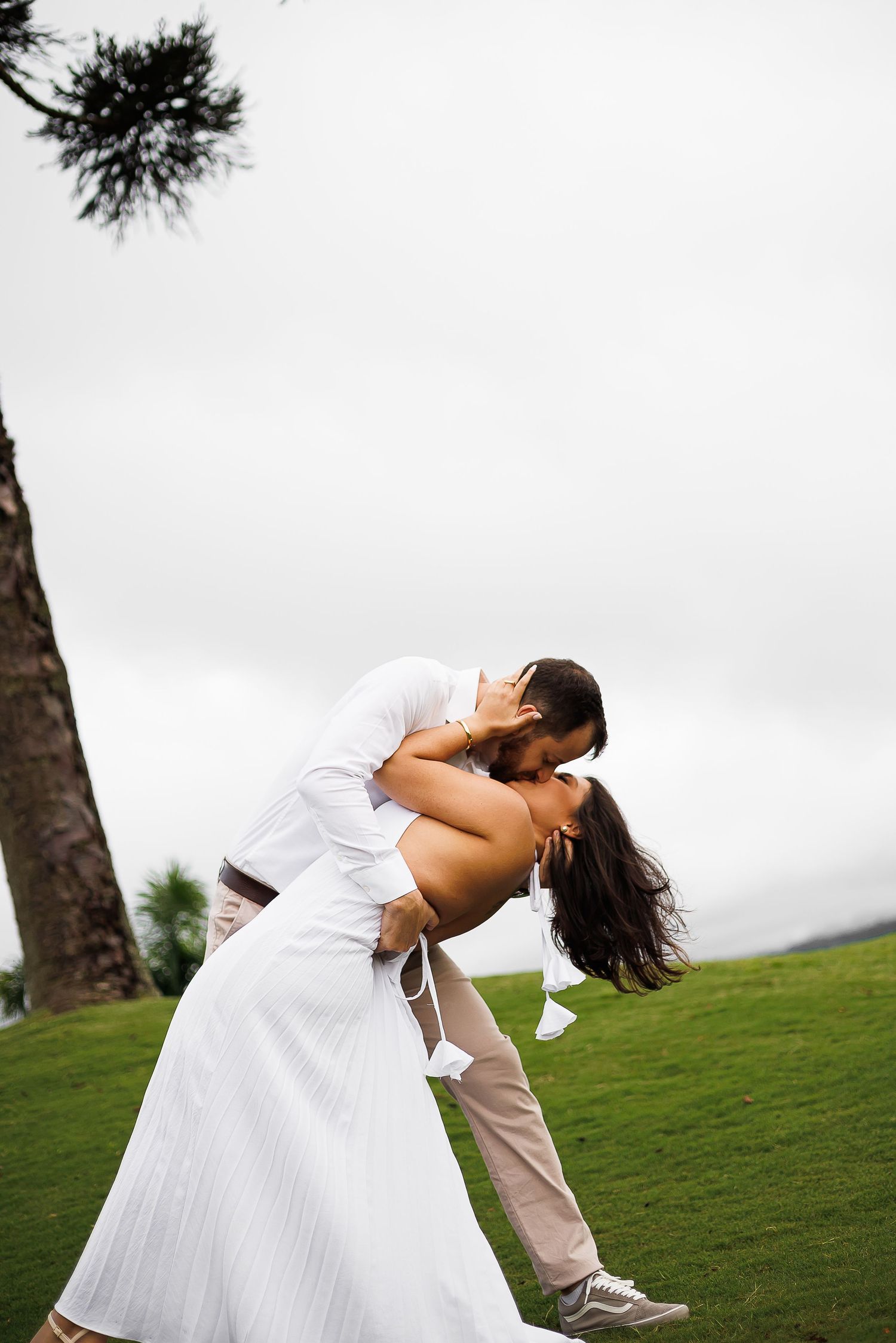 Couple sharing a romantic dip kiss on green lawn, bride in white dress, groom in casual attire, overcast sky.