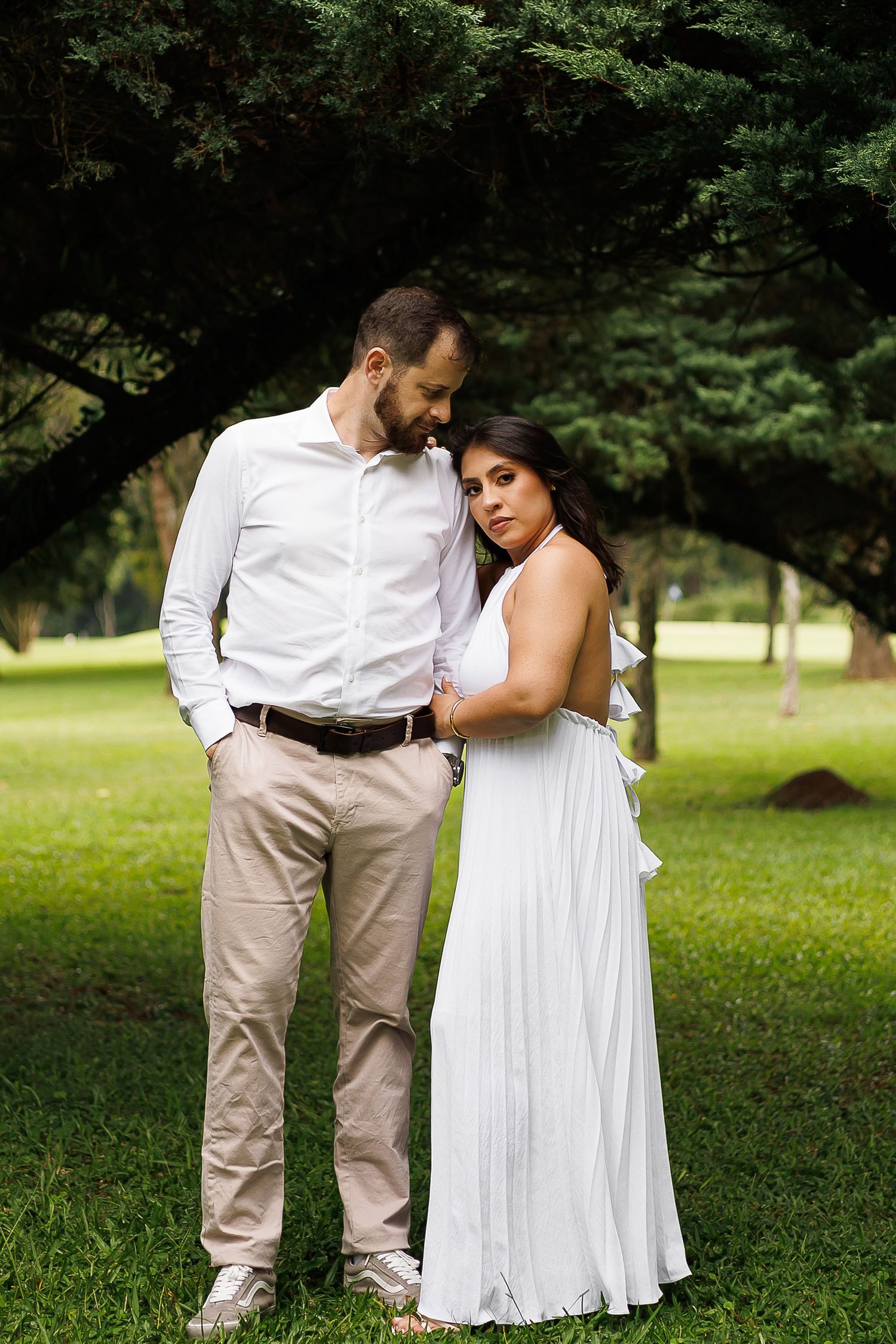 Couple posing in a lush green park, man in white shirt and khakis, woman in elegant white maxi dress.