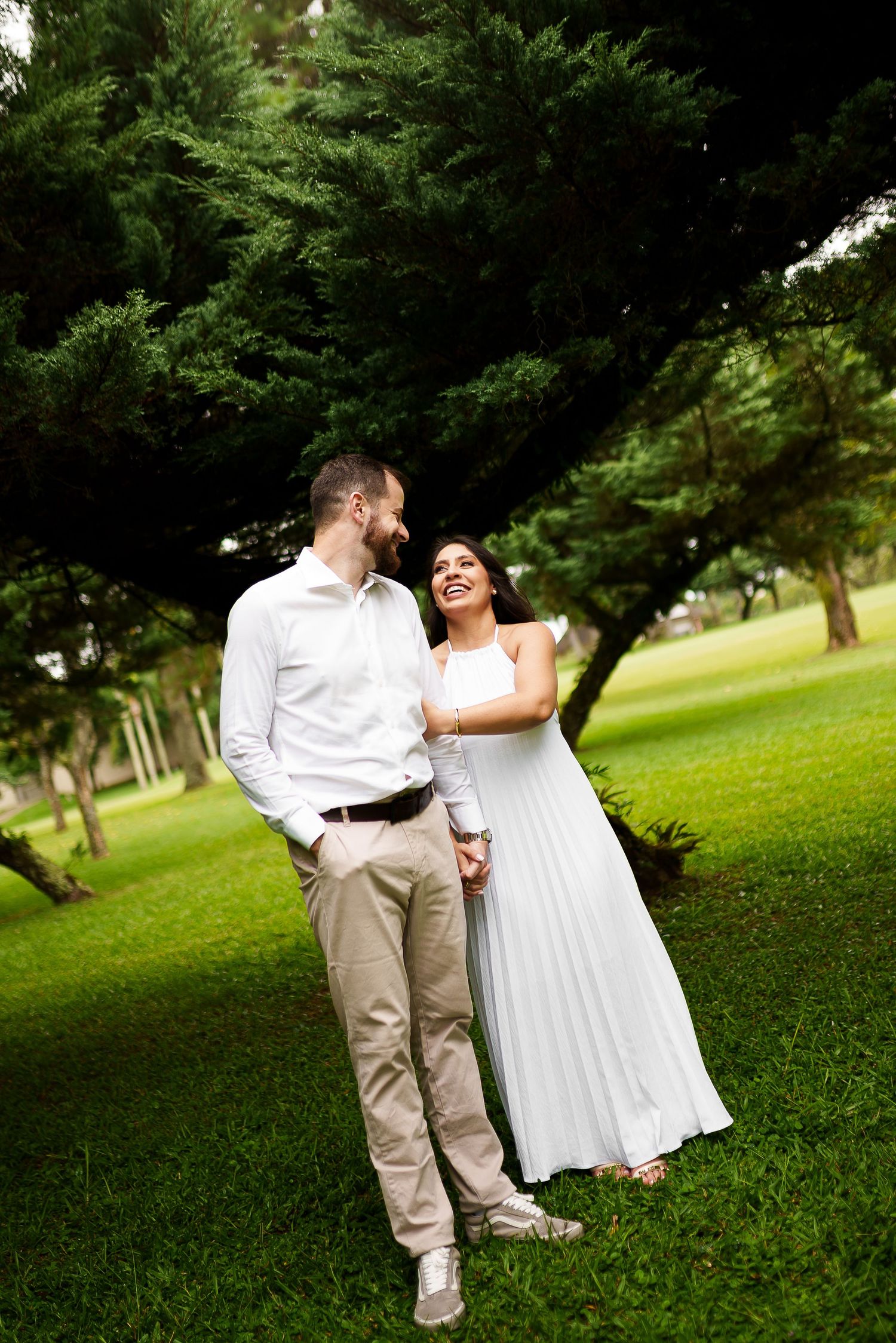 A happy couple in white outfits holding hands and laughing together in a lush green park setting.