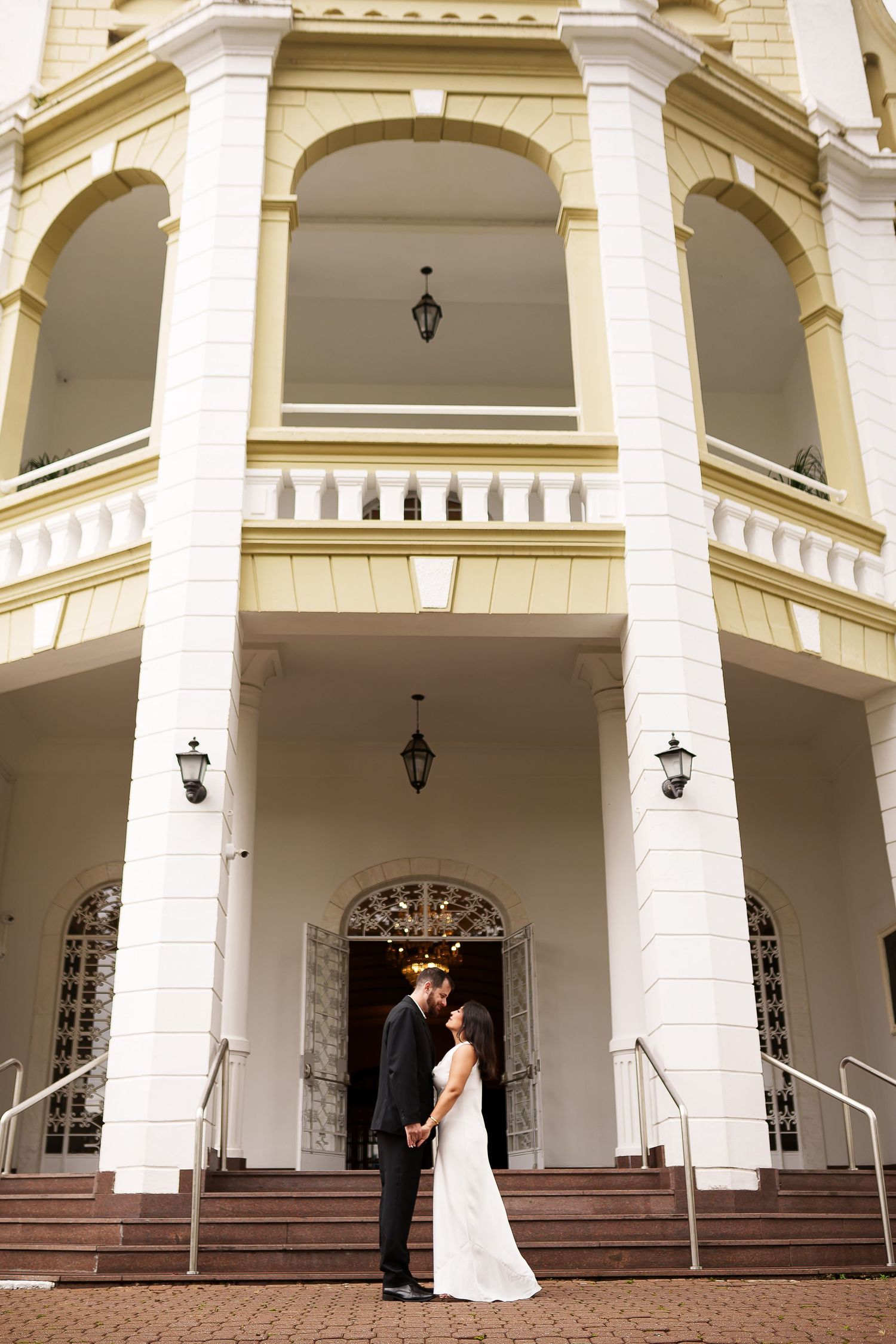 Bride and groom share a romantic moment on the steps of an elegant white colonial building with arched windows.