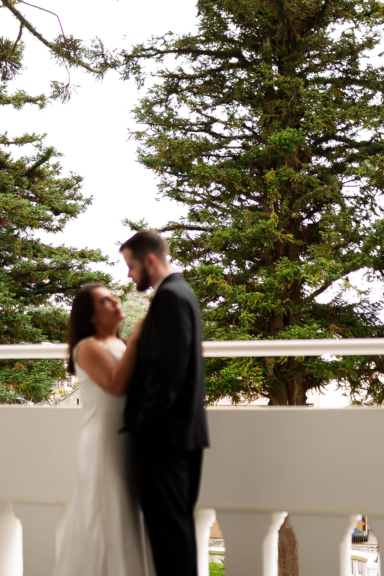 Blurred bride and groom share a romantic moment on a white balcony surrounded by lush green trees.