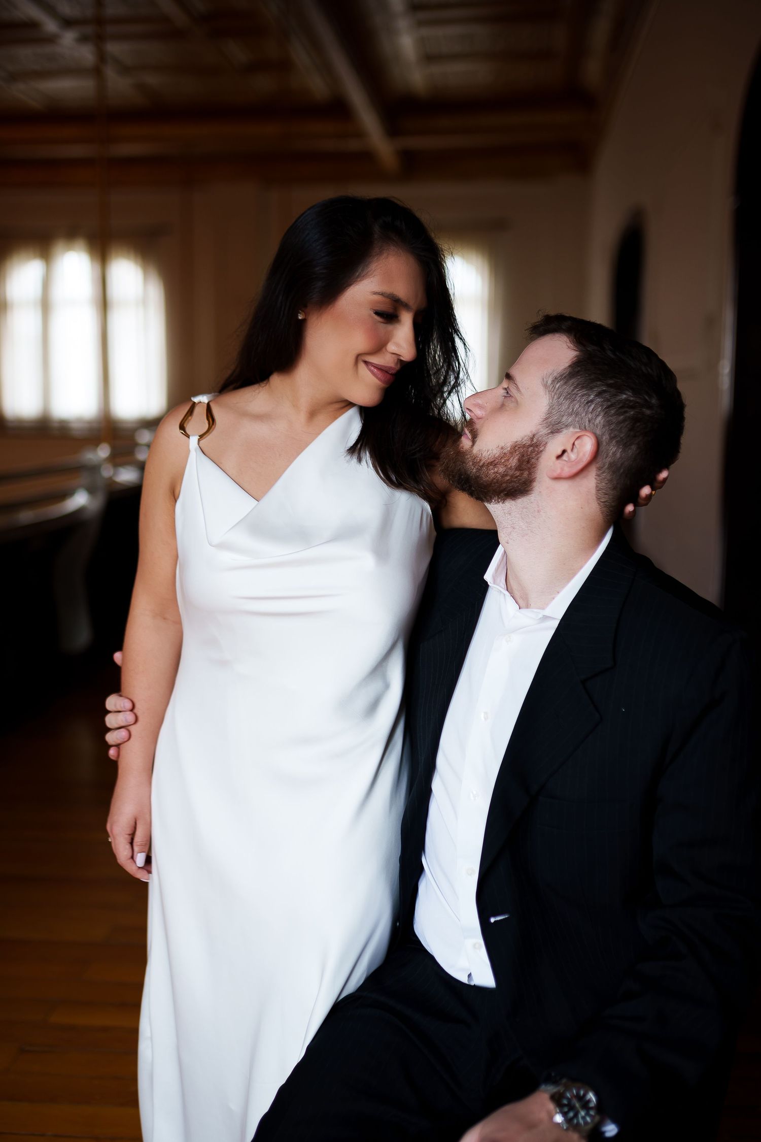 A bride in a white gown and groom in a black suit share a romantic gaze indoors.
