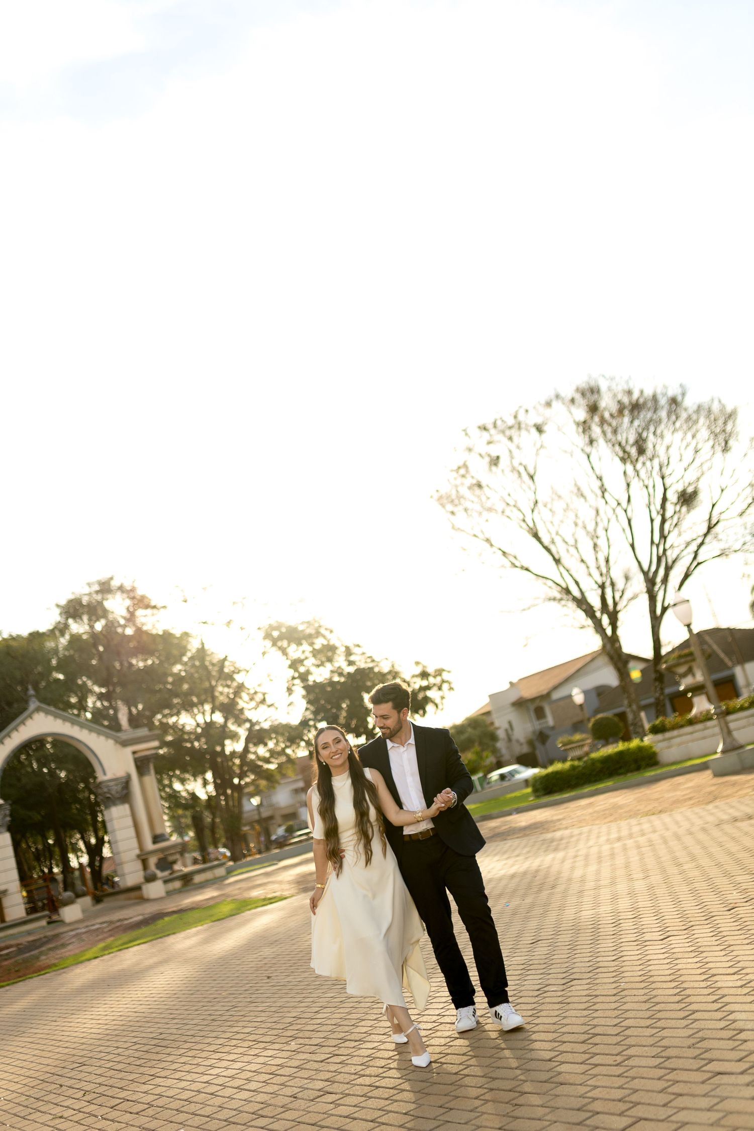 A couple in formal attire walks hand-in-hand on a brick plaza, bathed in warm golden sunlight with trees in the background.