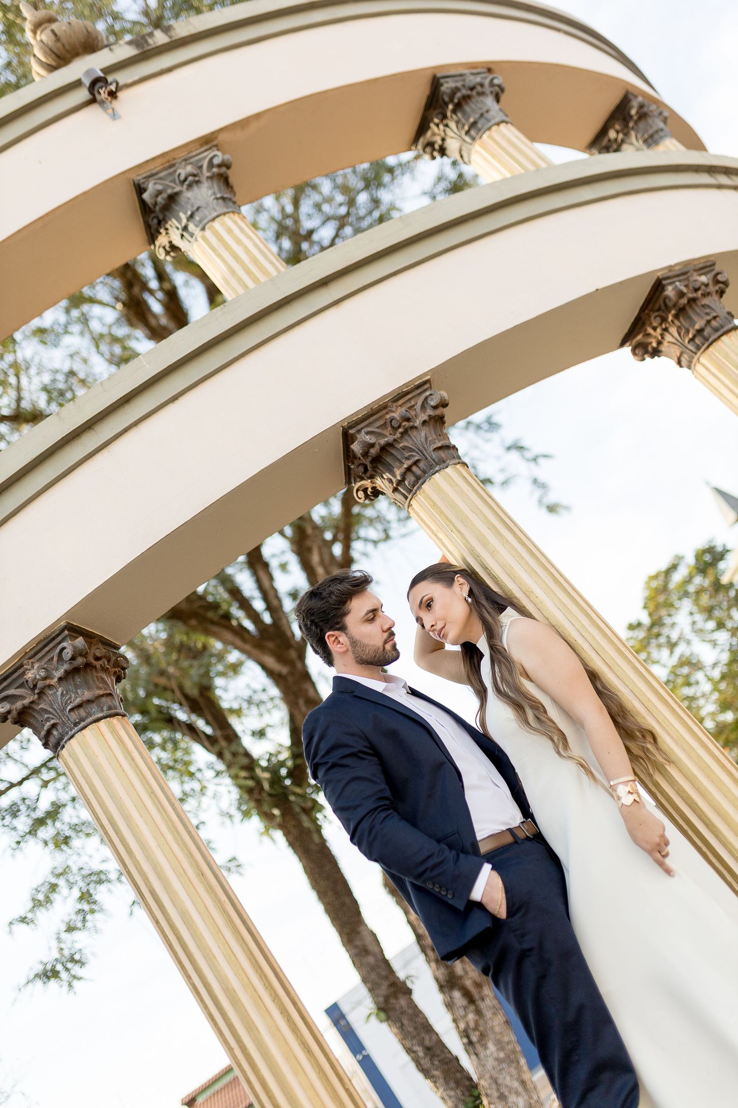 Elegant couple in wedding attire posing under ornate classical columns with arches outdoors.