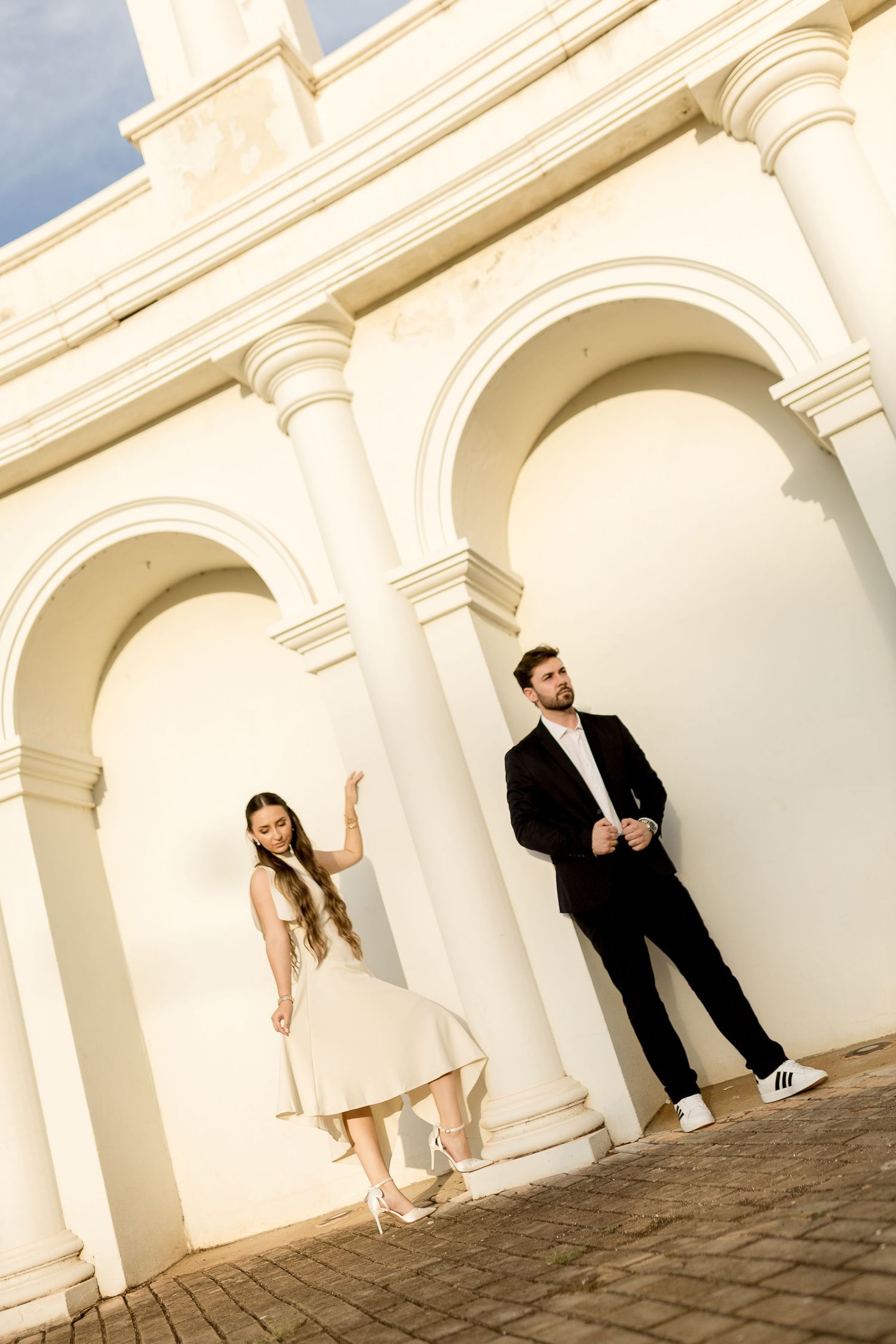 Elegant couple posing by white arched columns, woman in cream dress, man in black suit, classic architecture backdrop.