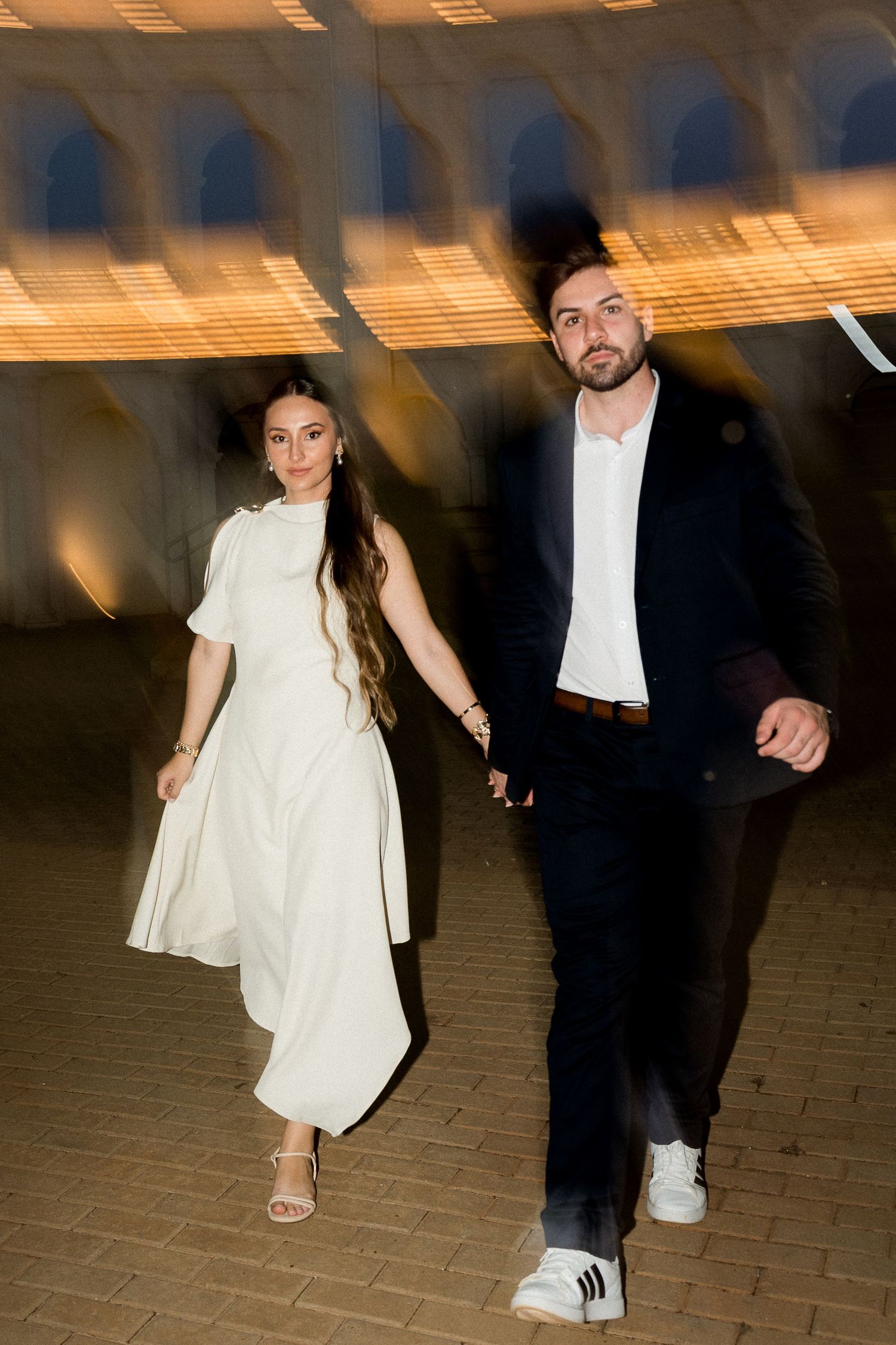 Couple walking hand in hand at night, woman in white dress, man in dark suit with white sneakers.