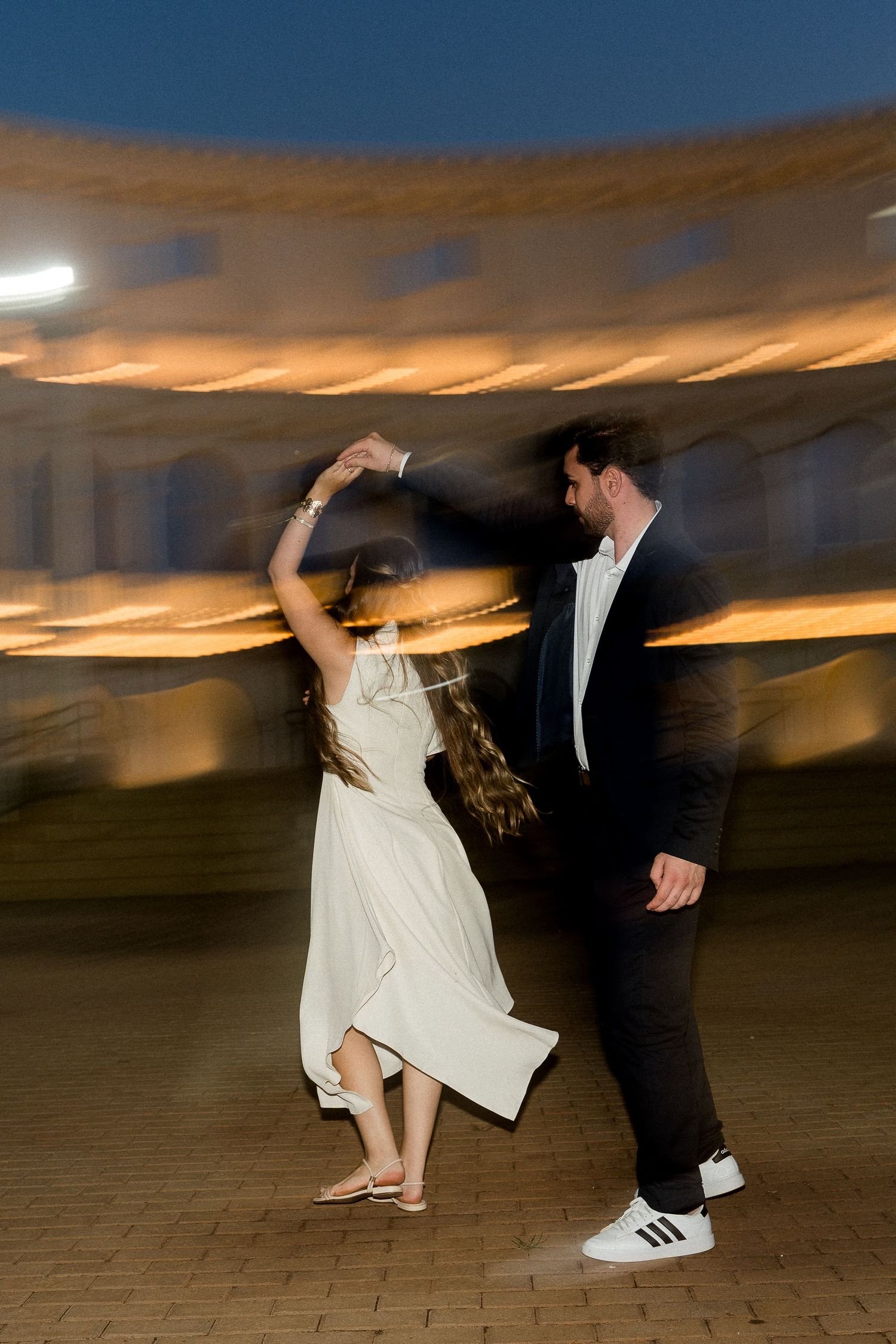 Couple dancing outdoors at night near illuminated building, woman in white dress, man in dark suit and Adidas sneakers.