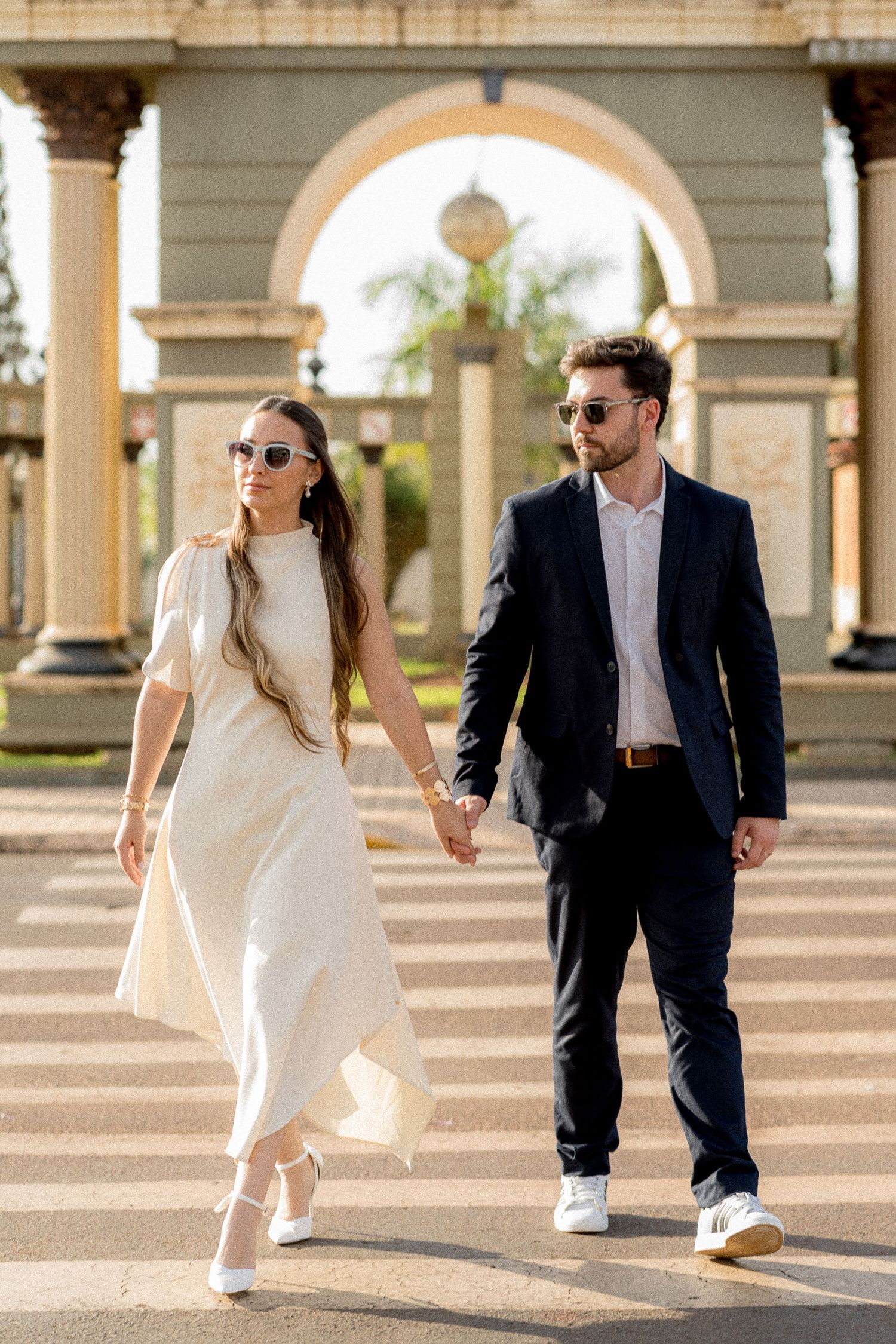 Elegant couple holding hands walking across a crosswalk, woman in white dress and man in navy suit, with ornate arch backdrop.