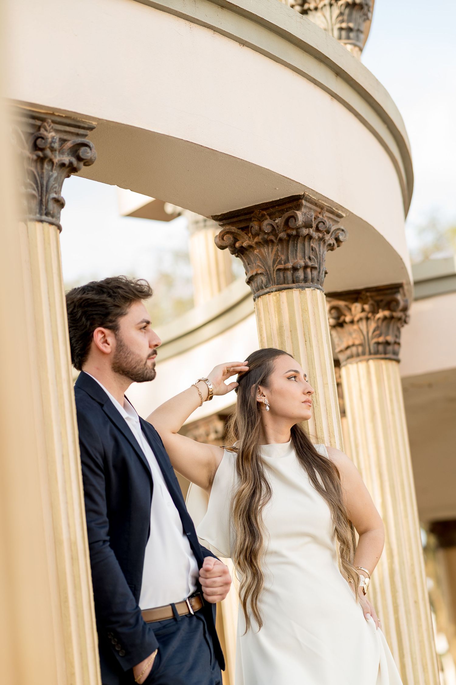 Elegant couple posing near ornate Corinthian columns, woman in white dress and man in navy blazer.