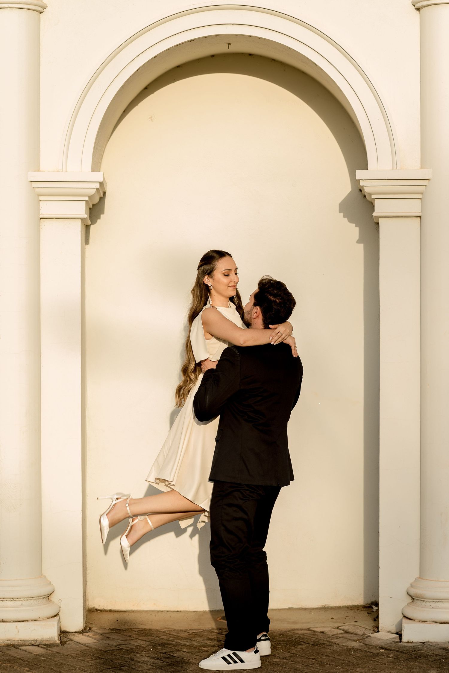 Groom lifting bride in white dress under elegant arch, romantic wedding portrait outdoors.