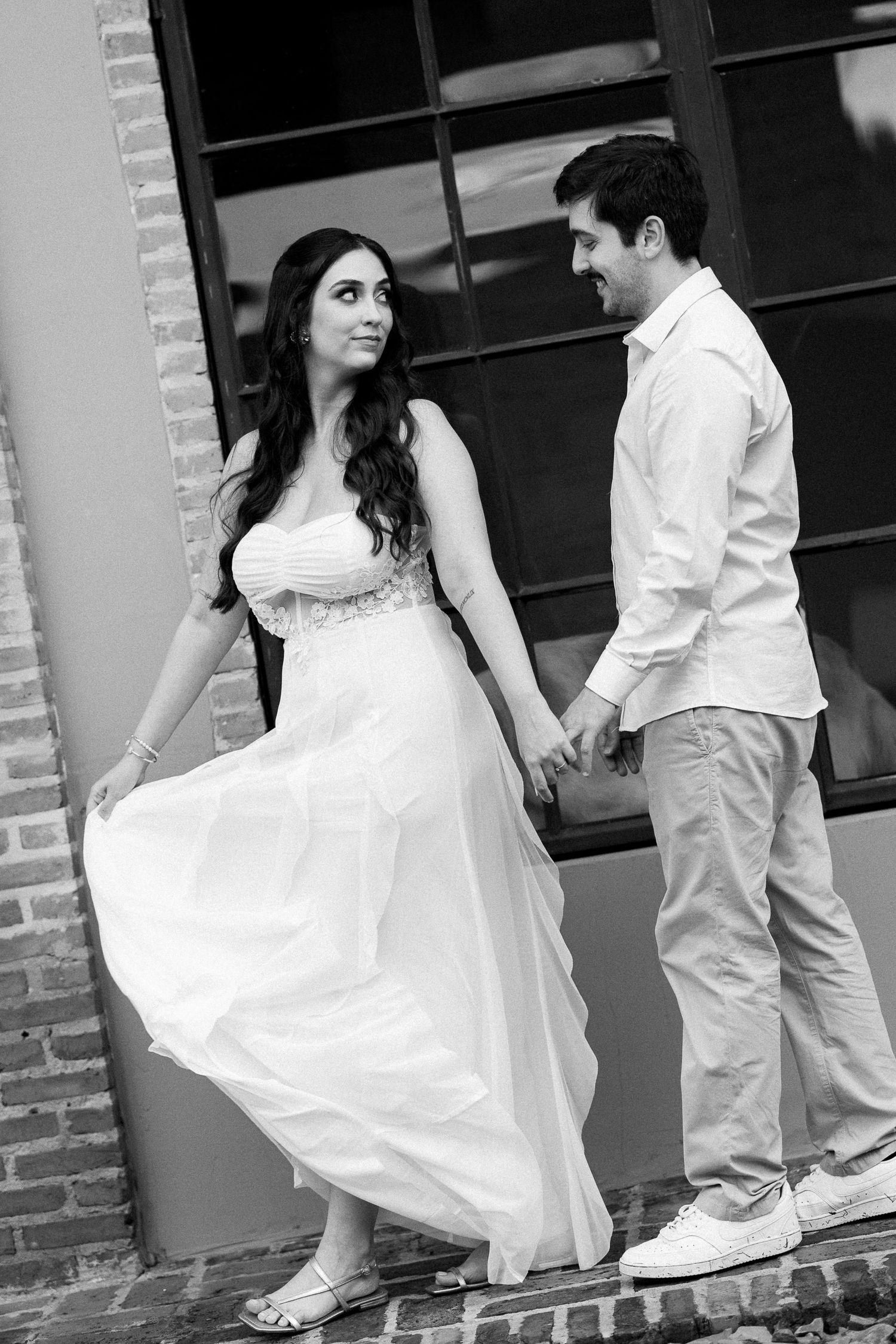 Black and white photo of a couple holding hands, woman in flowing white dress, man in casual attire, posing near brick building.