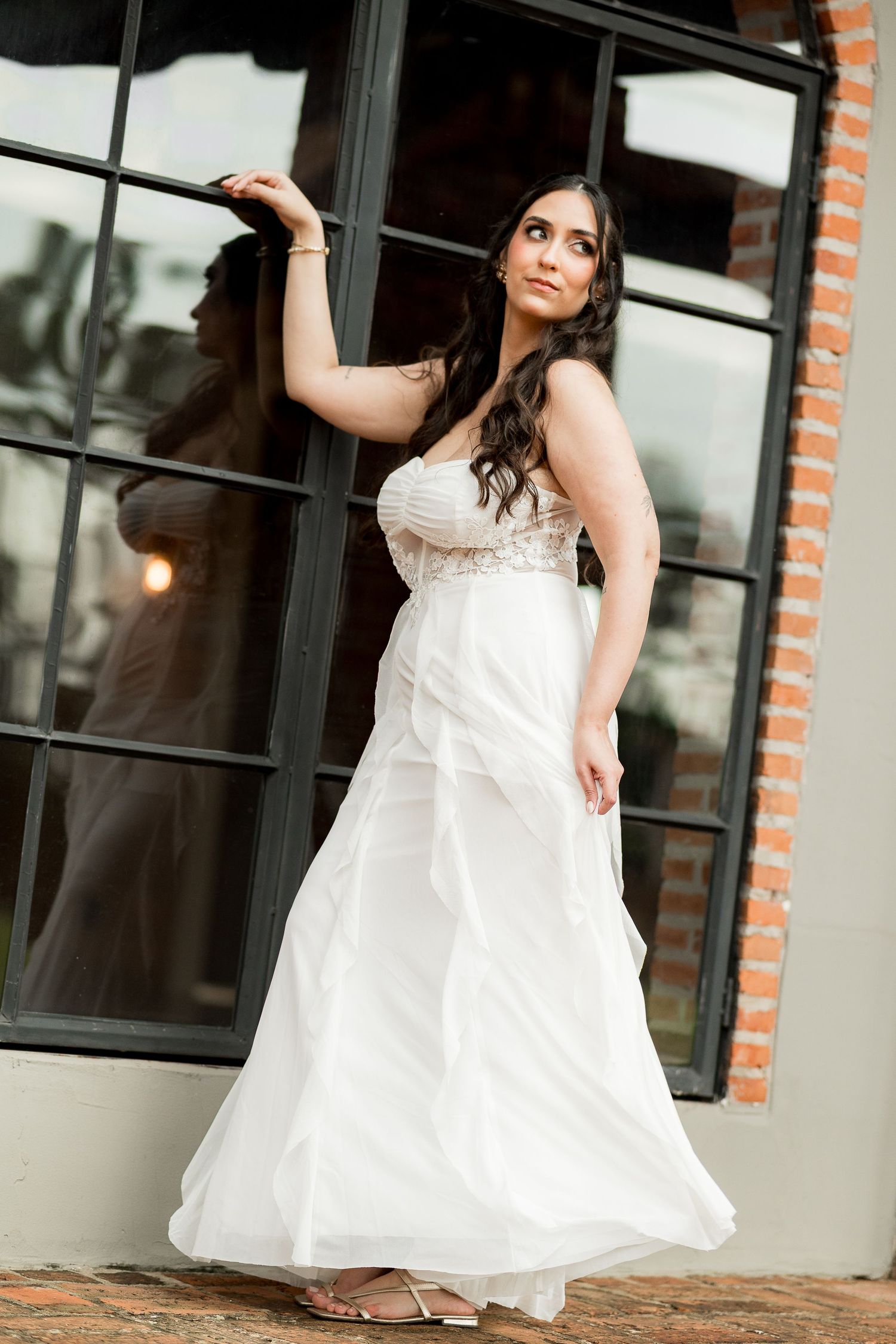 Bride in white strapless wedding gown posing elegantly by brick building with large black-framed windows.