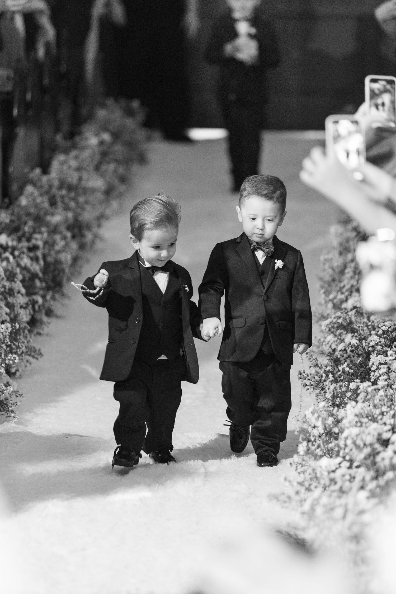 Two young ring bearers in matching black tuxedos walk down wedding aisle holding hands.