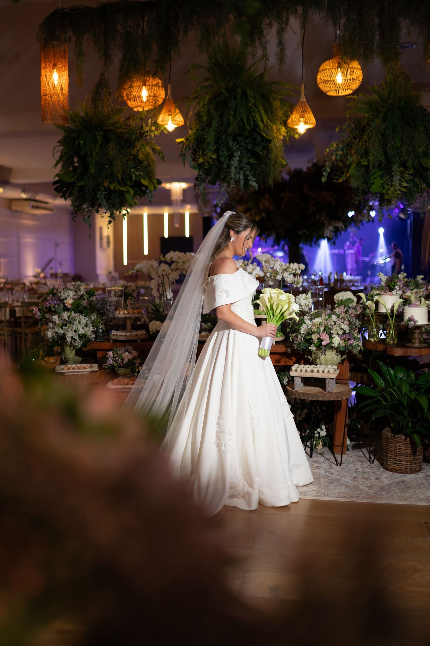 Bride in off-shoulder white gown with veil holds white bouquet inside a lush, warmly lit wedding venue.