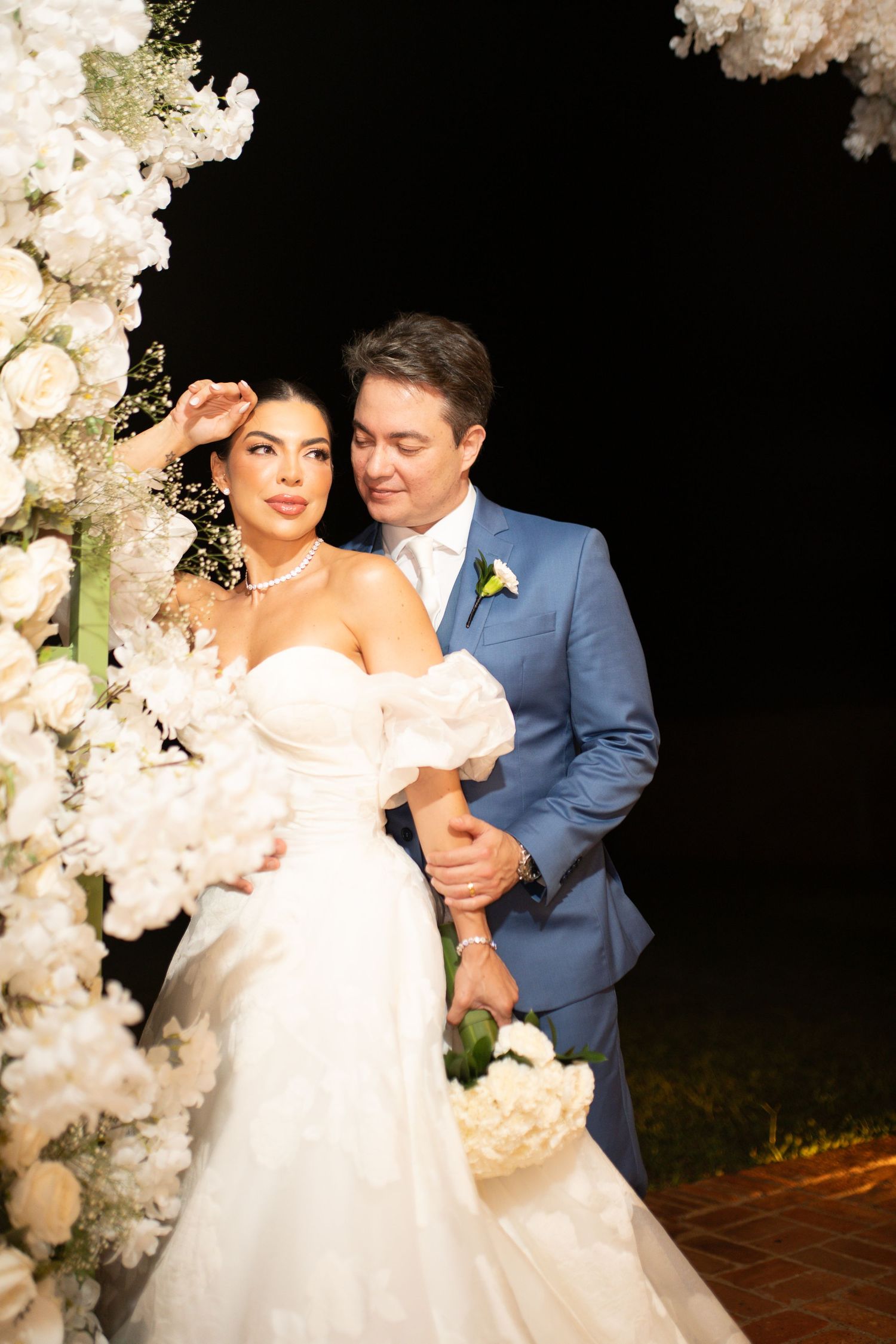 Bride in white strapless gown and groom in blue suit pose together under white floral arch at elegant evening wedding ceremony.