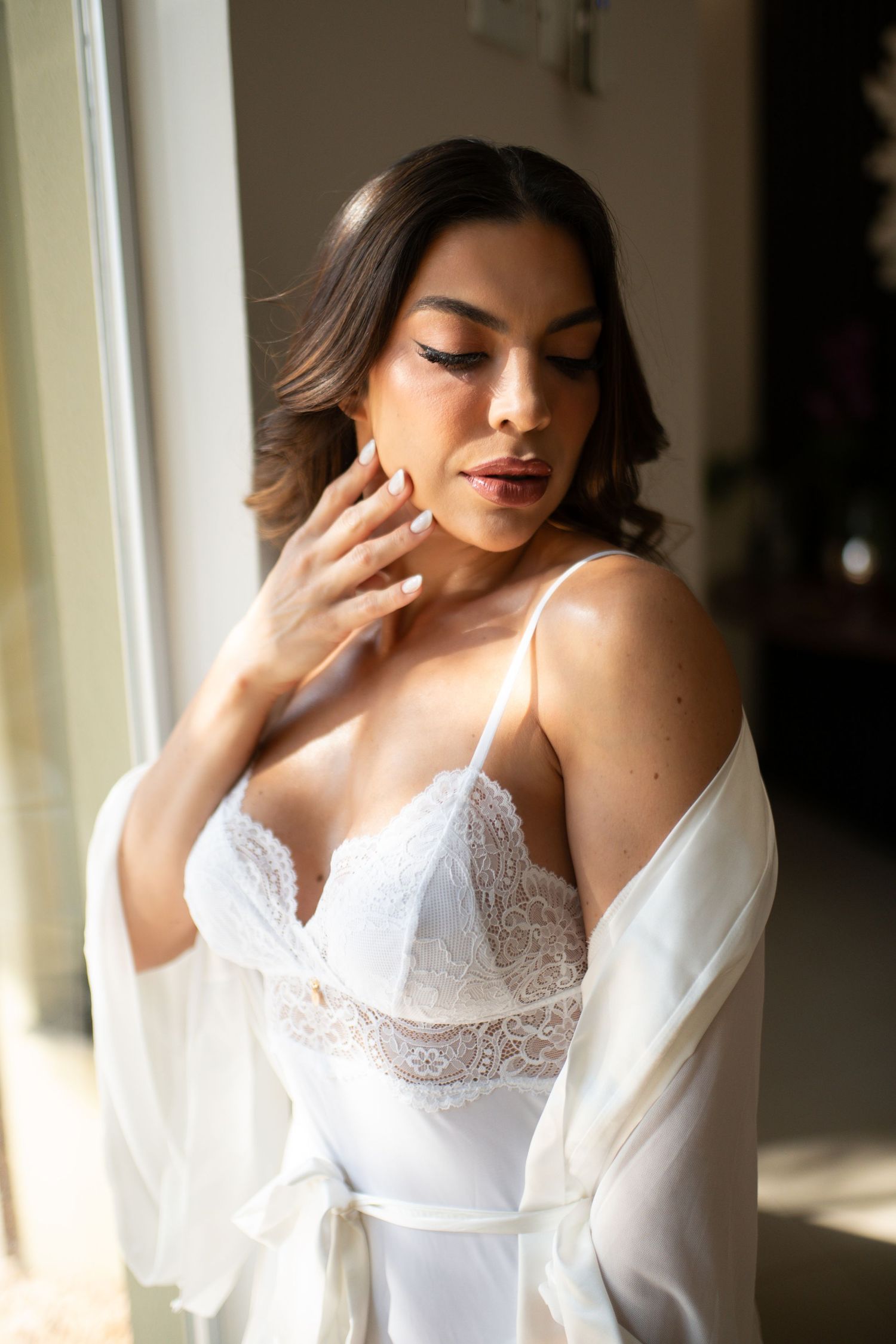 Woman in white lace lingerie and sheer robe posing elegantly near a sunlit window with soft glam makeup.