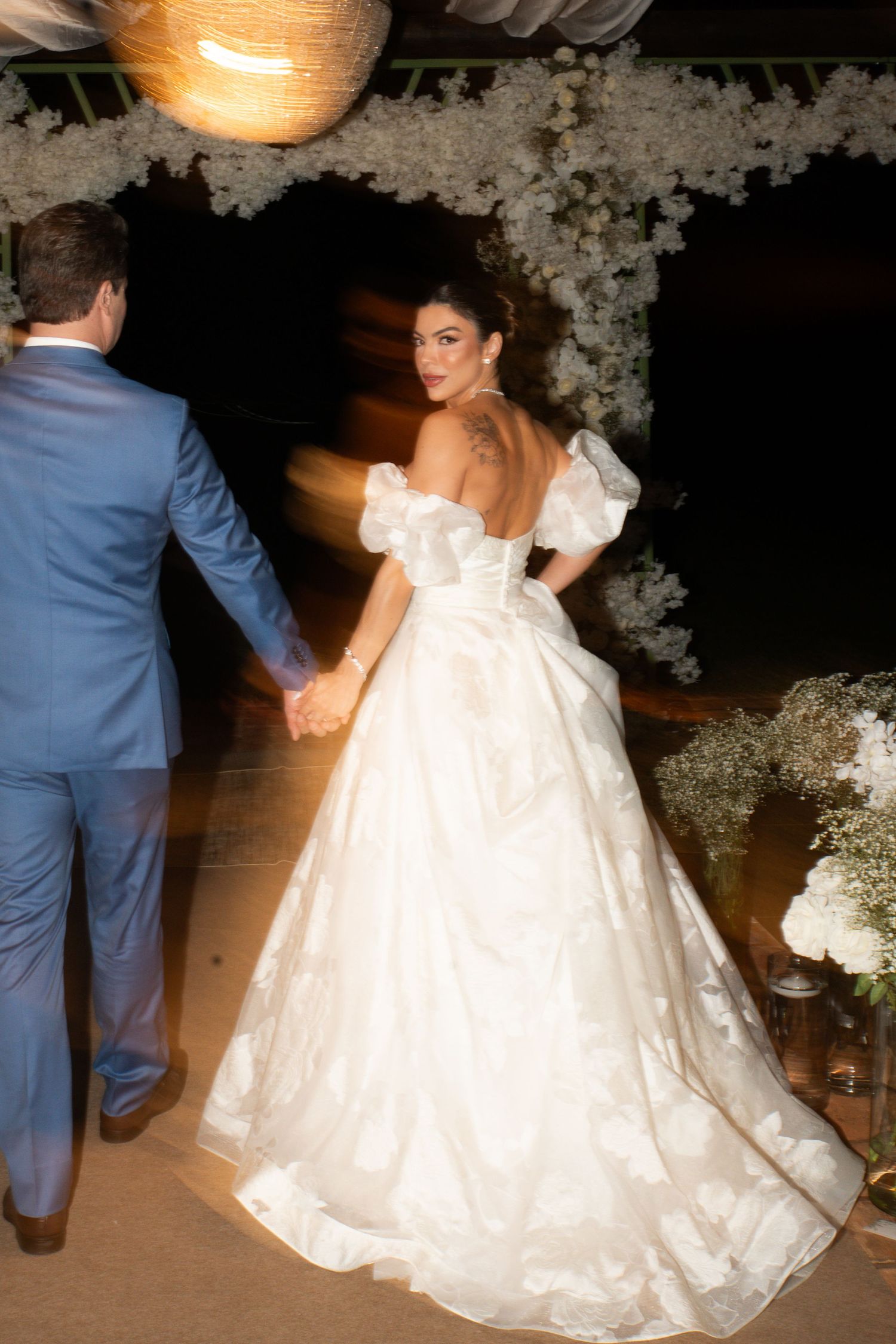 Bride in white off-shoulder ball gown holds groom's hand at elegant outdoor wedding reception with floral decor.