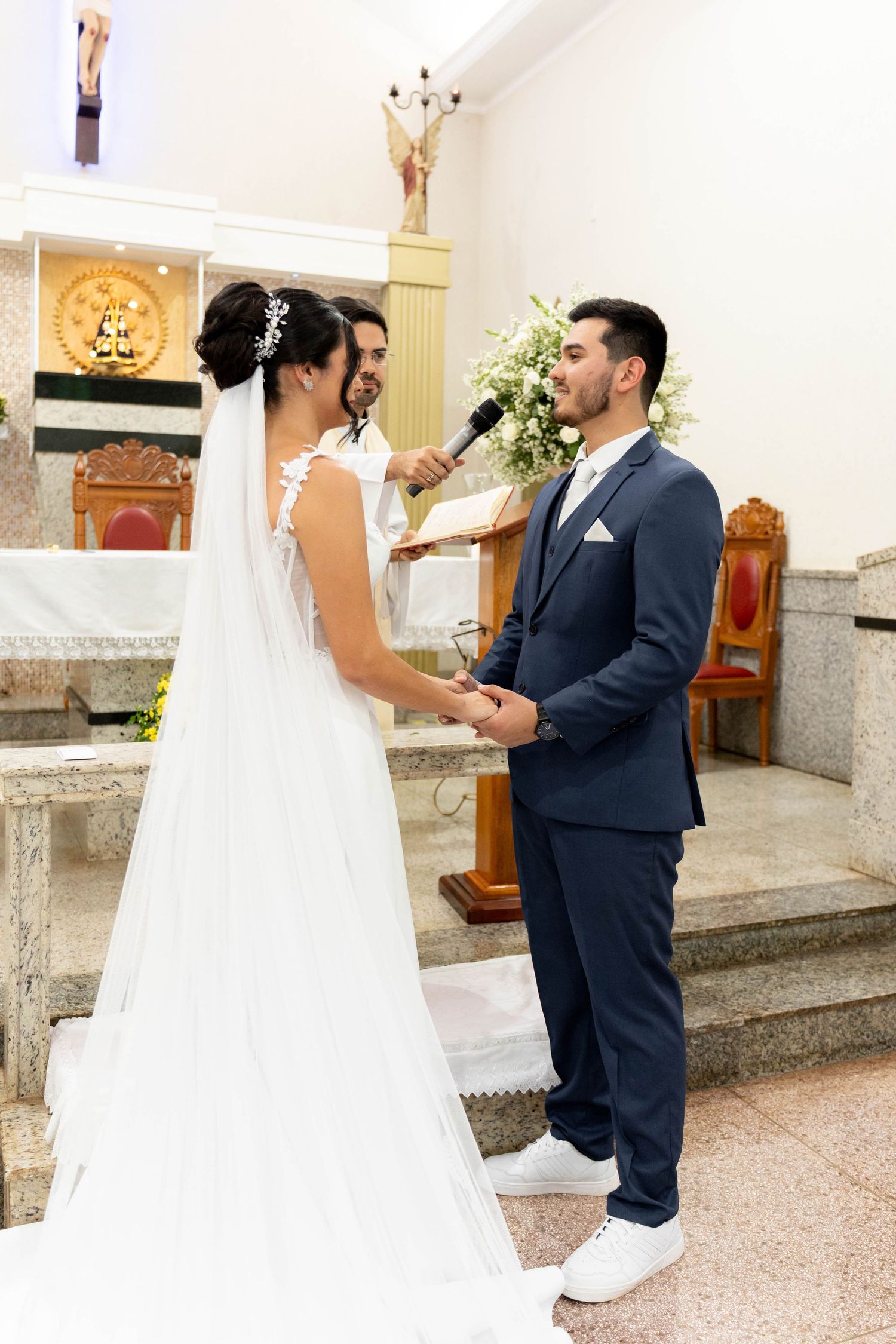 Bride and groom exchange vows at church altar, officiant holds microphone, bride in white gown with veil, groom in navy suit.