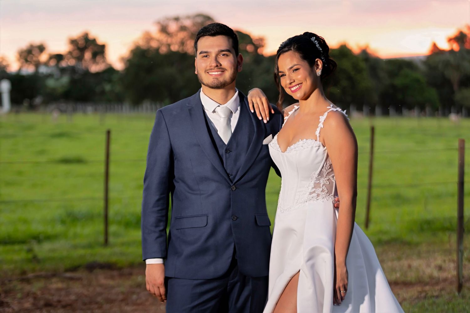 A couple in wedding attire poses at sunset with green fields behind them, the groom in a blue suit and bride in a white gown.
