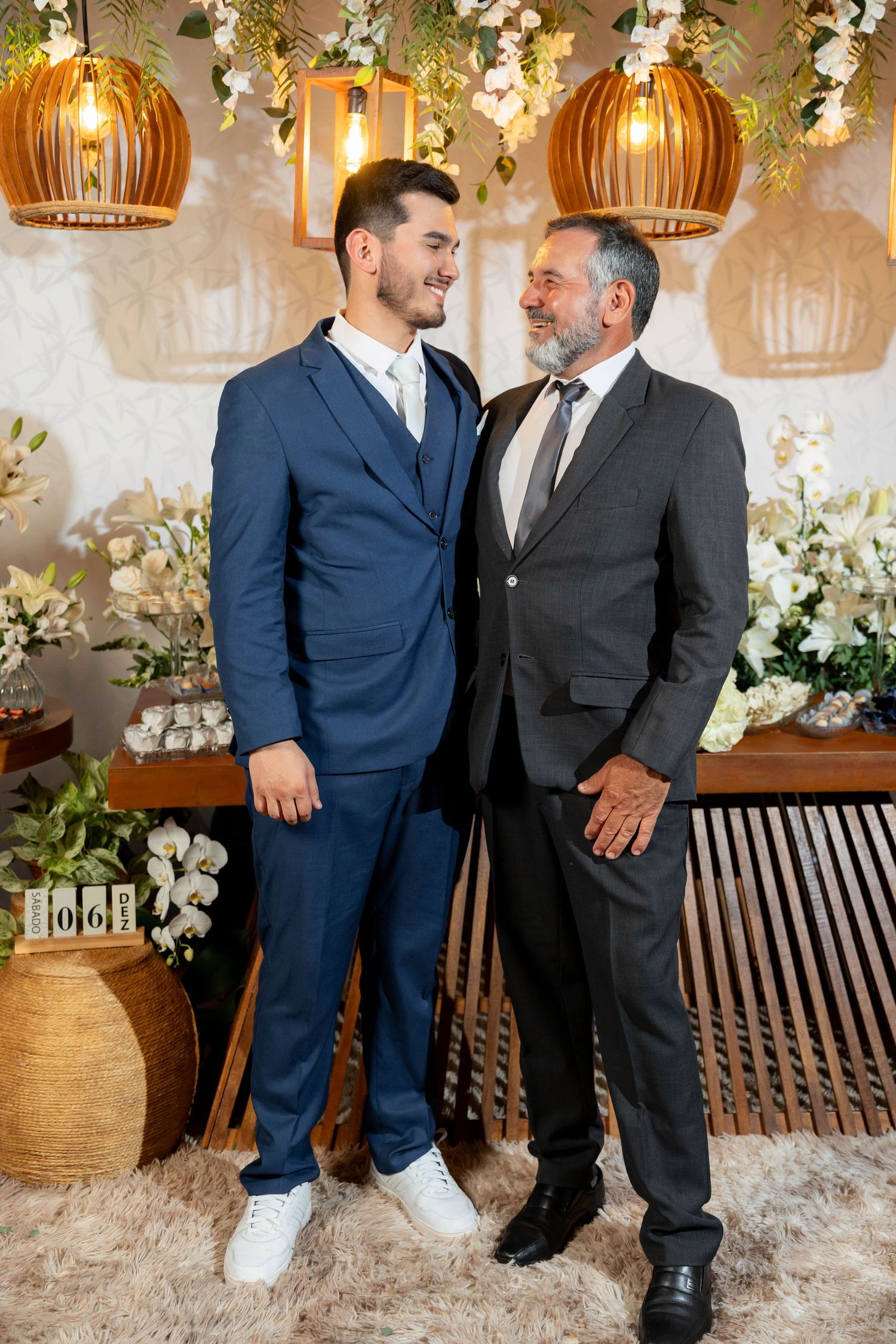 Two men in suits smiling at each other at a decorated event venue with floral arrangements and warm pendant lights.