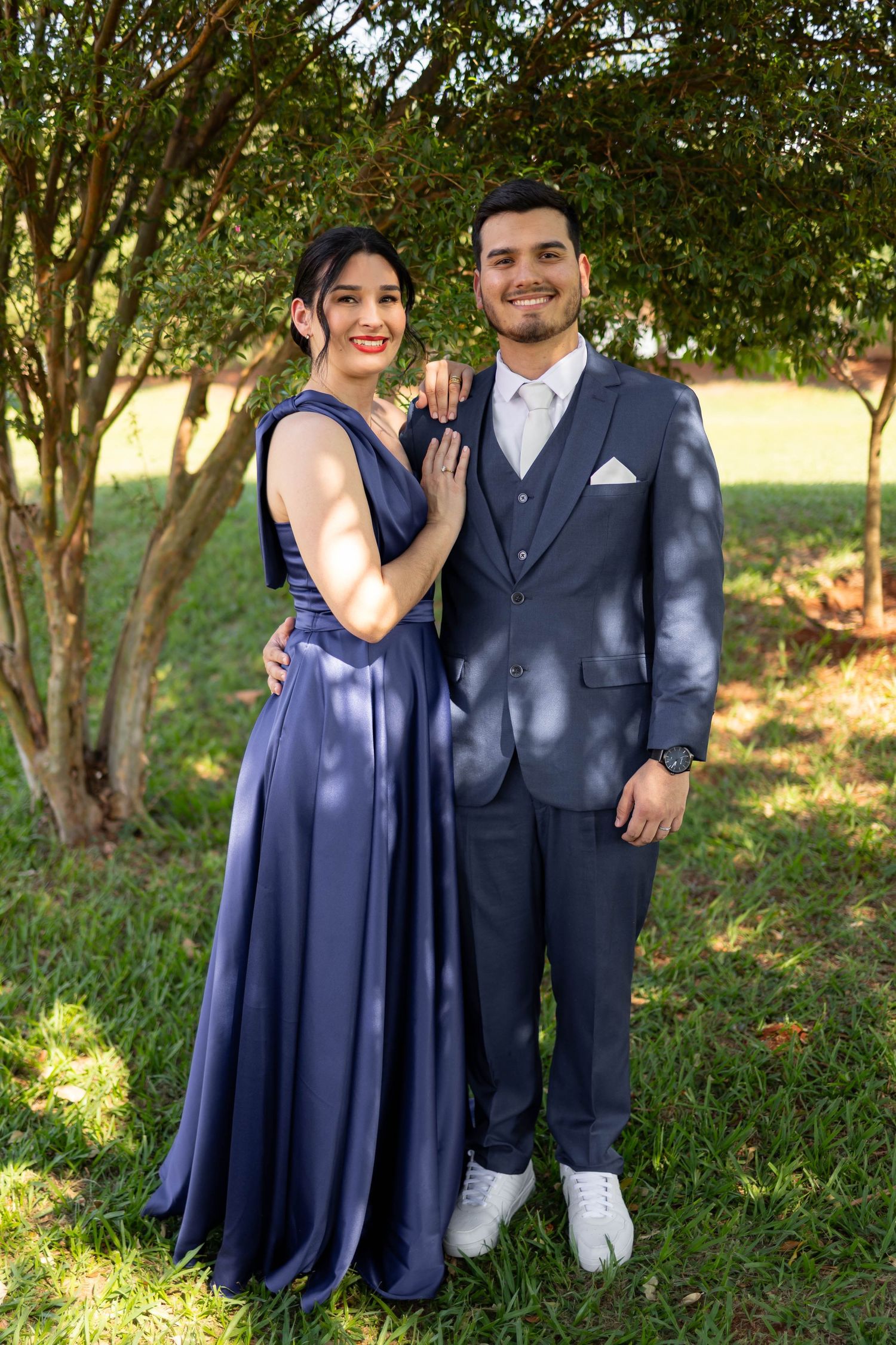 A couple in coordinating navy blue formal wear poses outdoors under a tree on a sunny day.