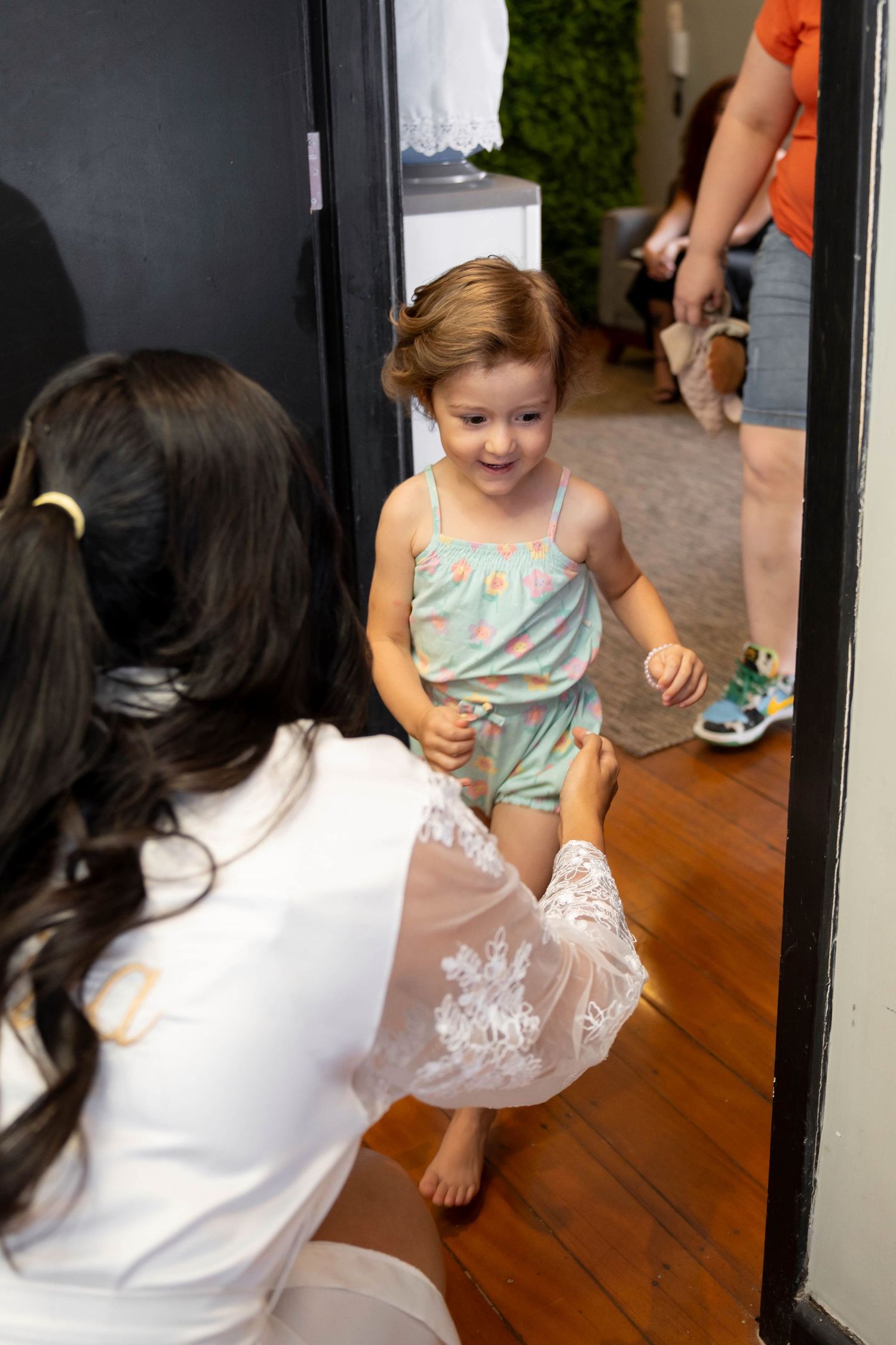A young girl in a mint romper smiles as a dark-haired woman kneels to help her with a lace skirt indoors.