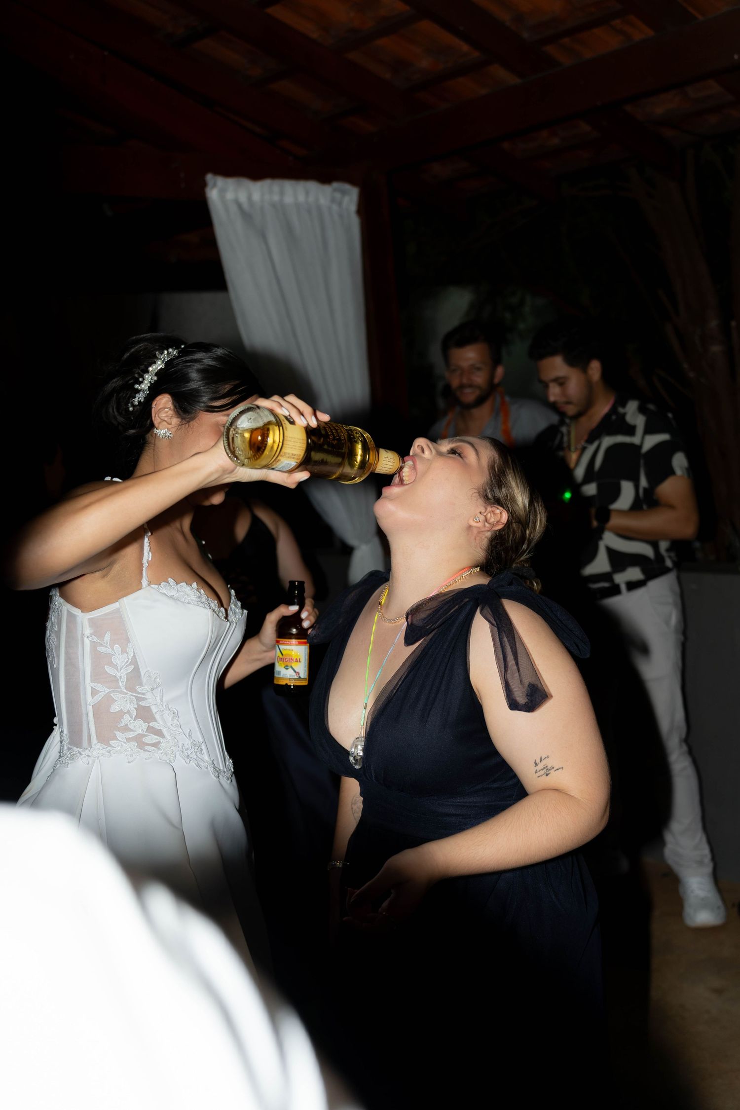Bride in white dress and bridesmaid in navy gown drinking from bottles at a lively wedding reception party.