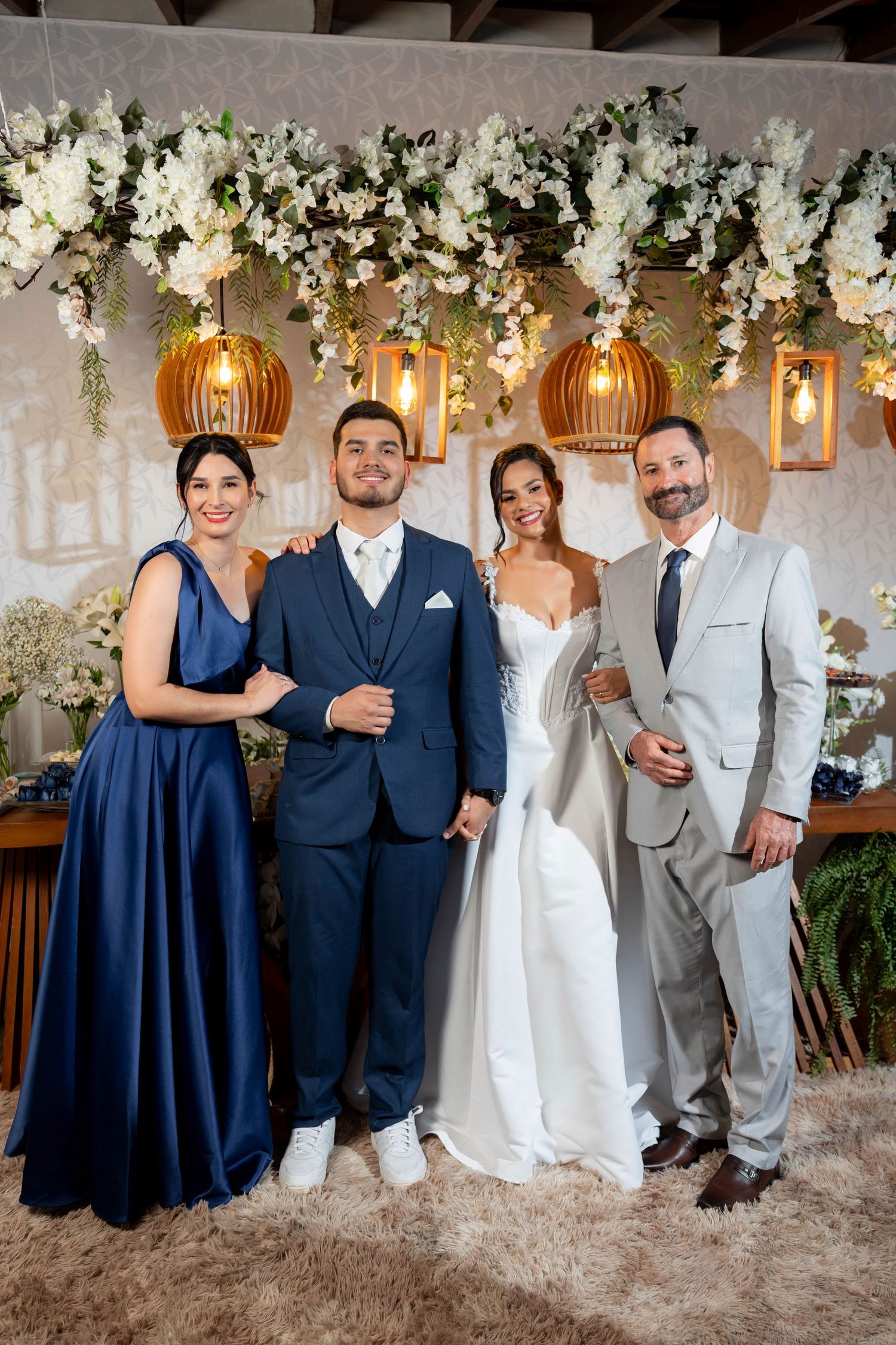 Wedding party of four posing elegantly before floral backdrop with hanging lanterns, bride in white gown, groom in navy suit.