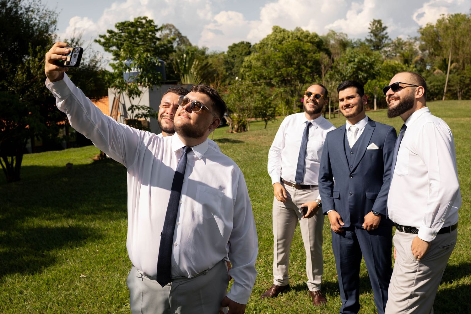 Groomsmen in suits taking a selfie outdoors on a sunny day at a wedding celebration.