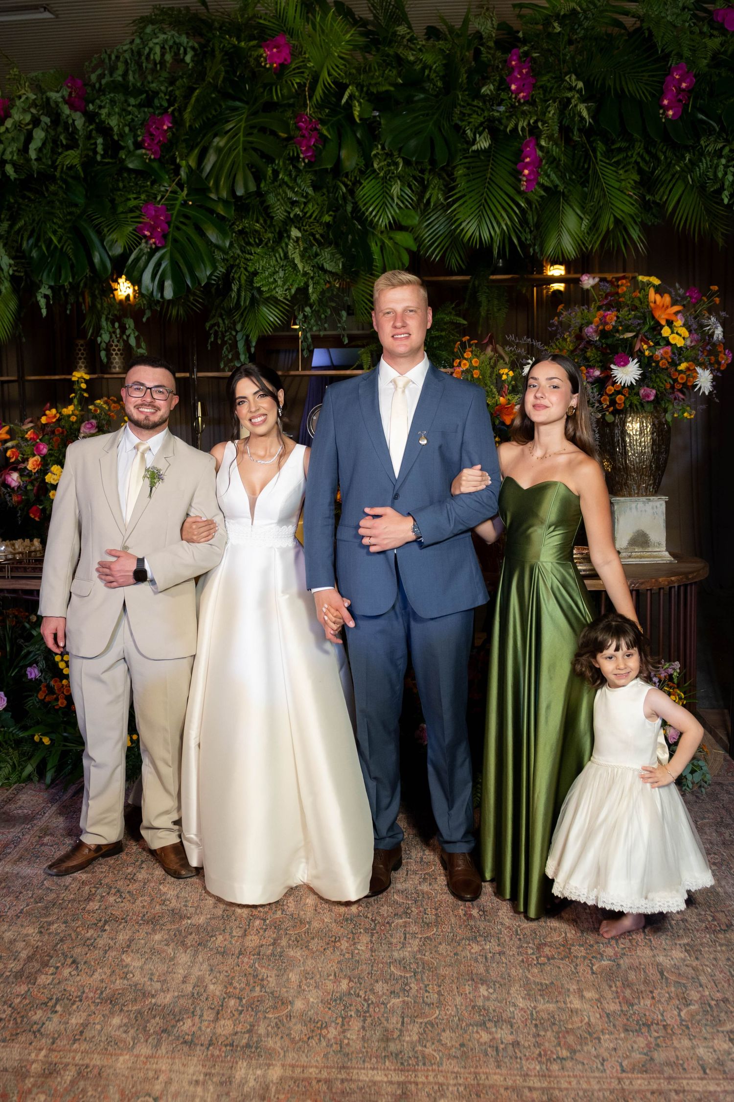 Wedding party poses with bride in white gown, groom in blue suit, bridesmaid in green dress, groomsman, and flower girl.
