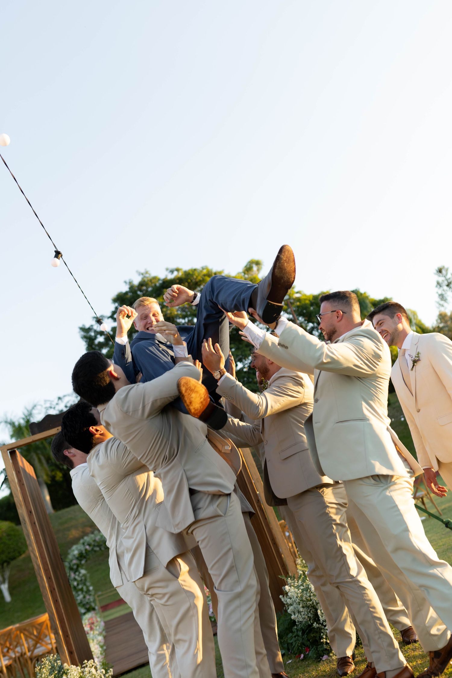 Groomsmen lifting the groom in the air outdoors at a wedding celebration under a clear blue sky.