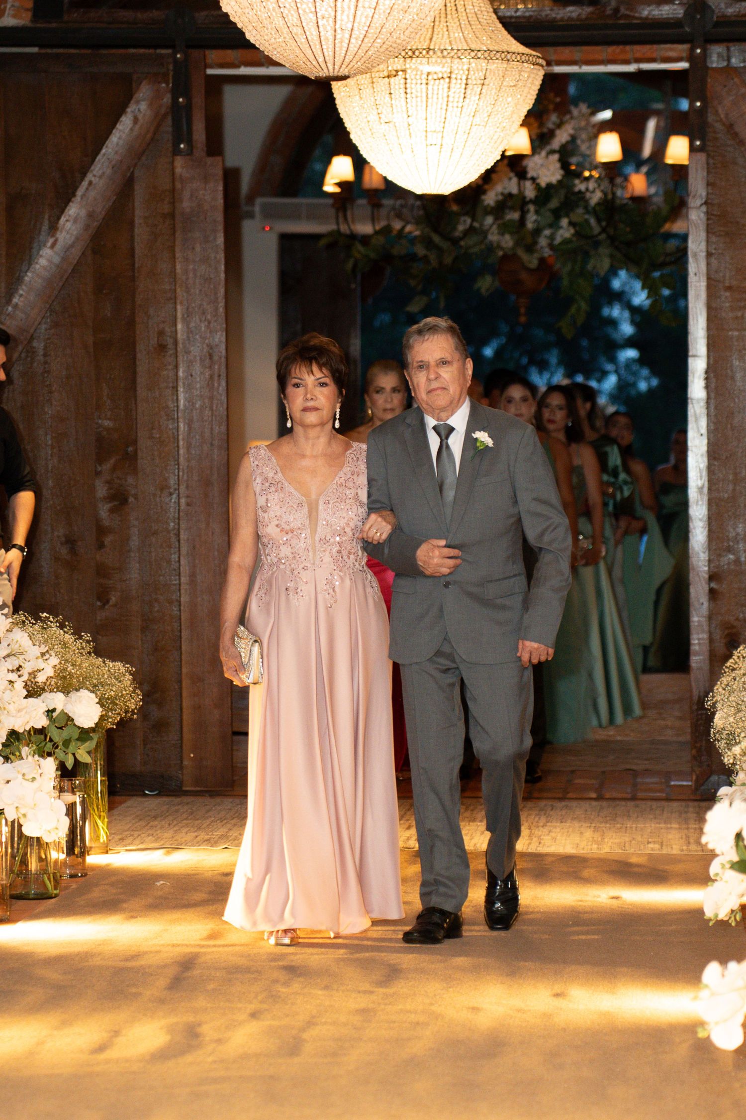 Elegant couple walking down wedding aisle, woman in pink gown and man in gray suit, rustic barn venue with floral decor.