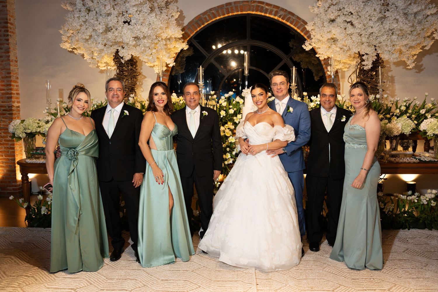 Wedding party poses together, bride in white gown, bridesmaids in sage green dresses, floral arch backdrop.