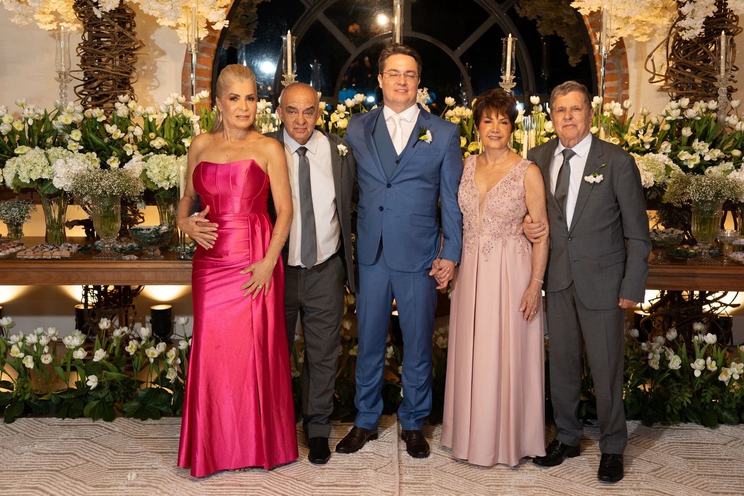 Five elegantly dressed people posing together at a formal wedding reception with floral decorations.