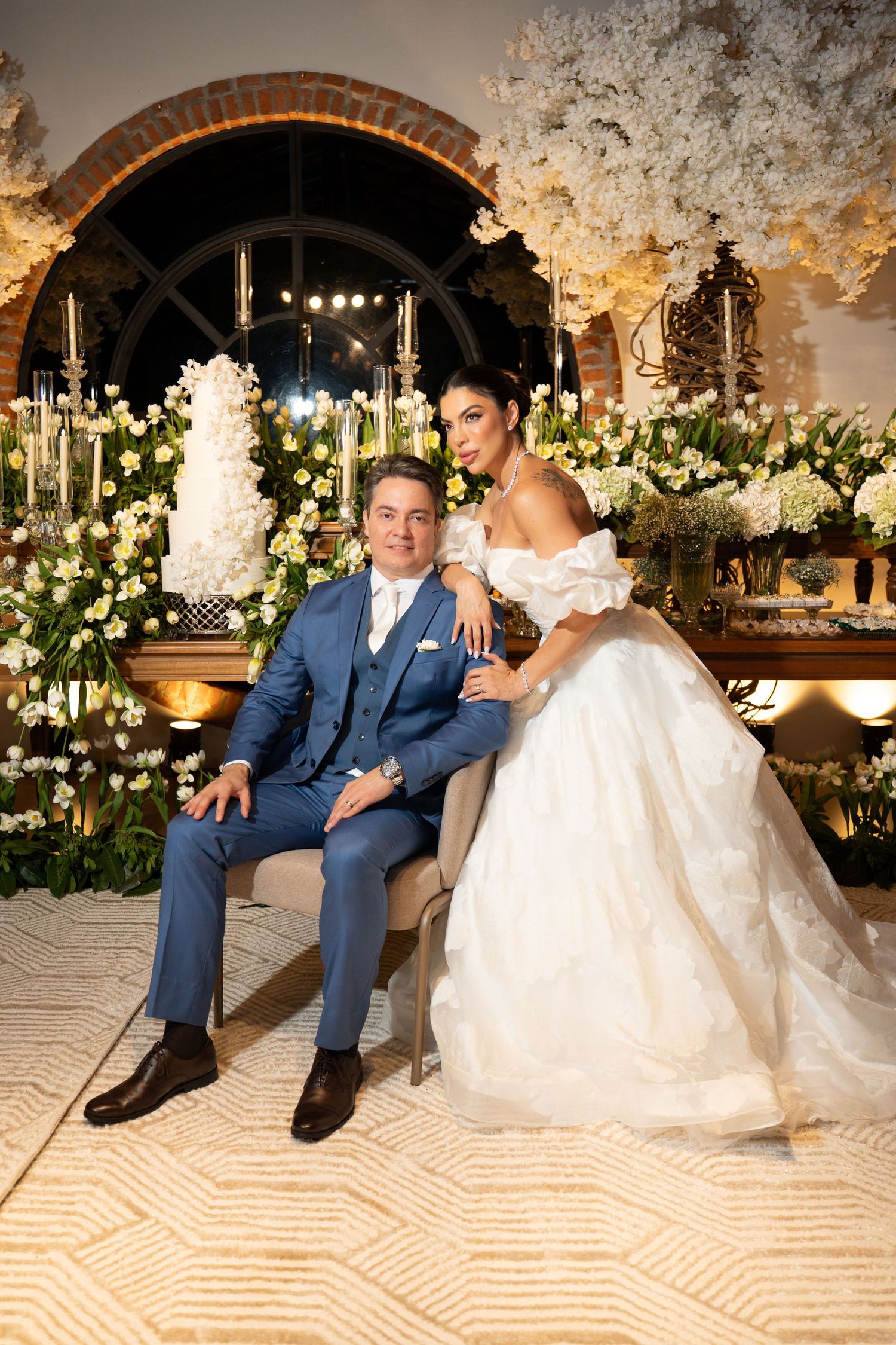 Elegant wedding couple posing before a floral backdrop, groom in blue suit seated, bride in white ballgown beside him.