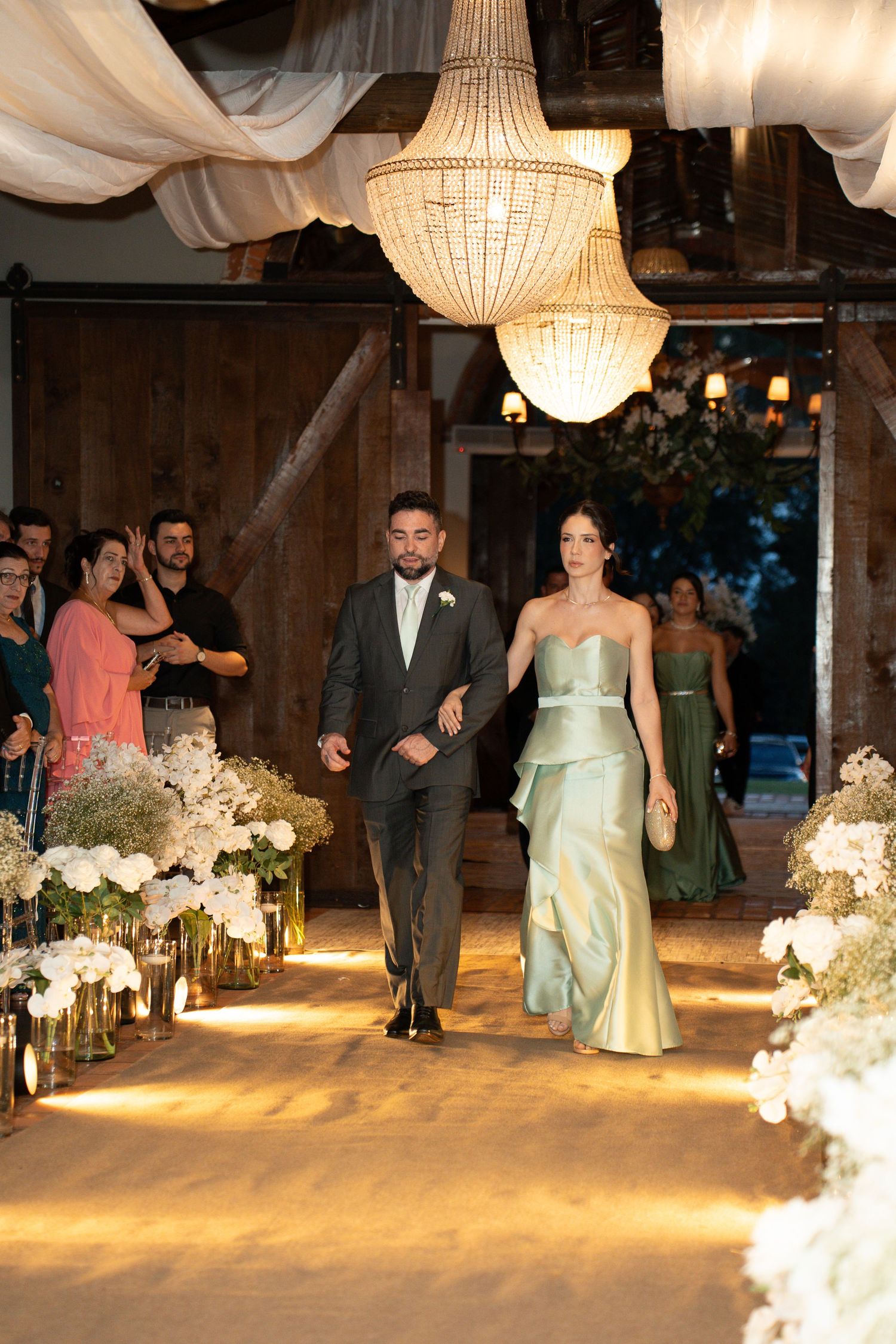 Elegant wedding procession in a barn venue with chandeliers, floral arrangements, and guests as a couple walks down the aisle.