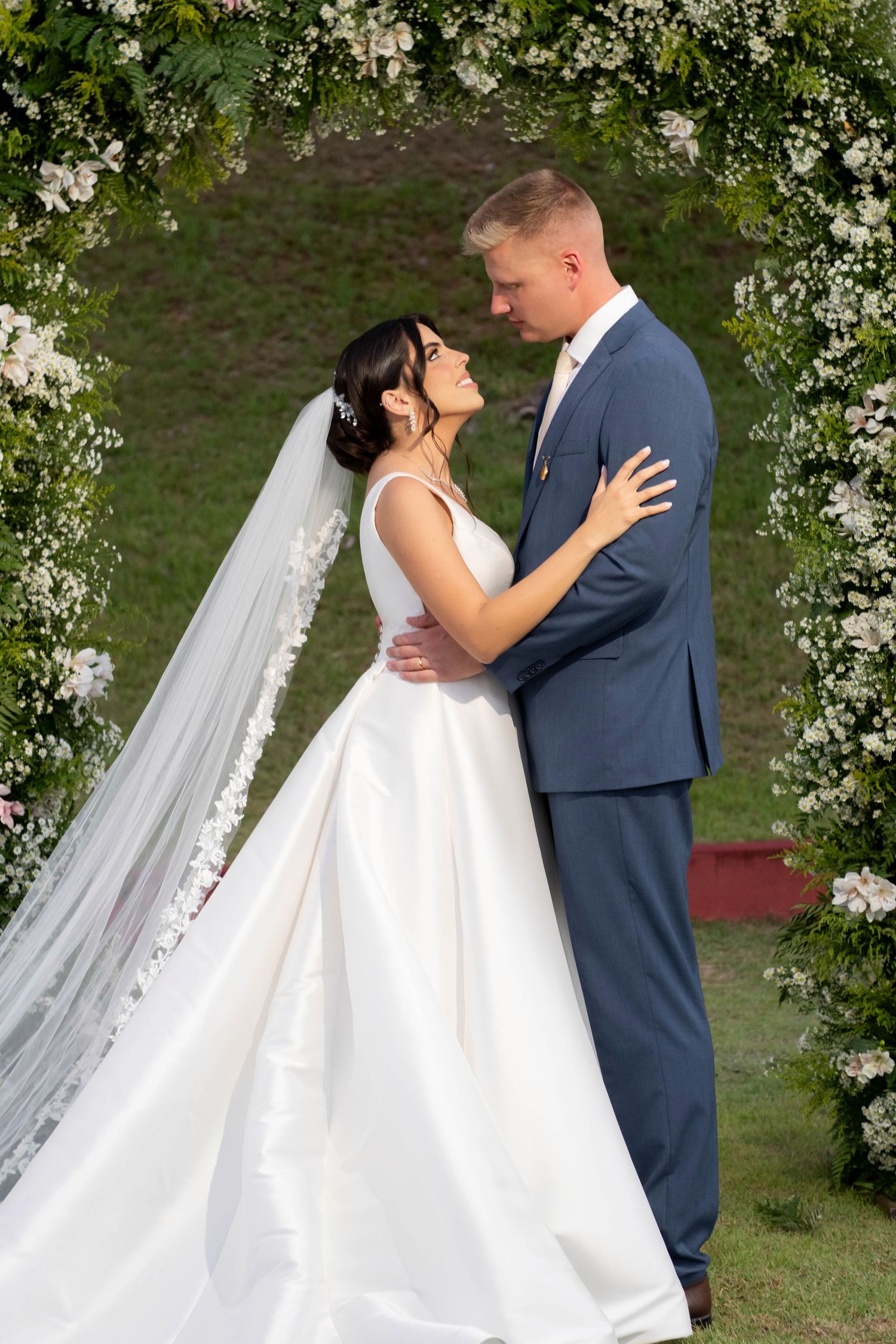 Bride in white gown and veil embraces groom in blue suit before floral arch outdoors.