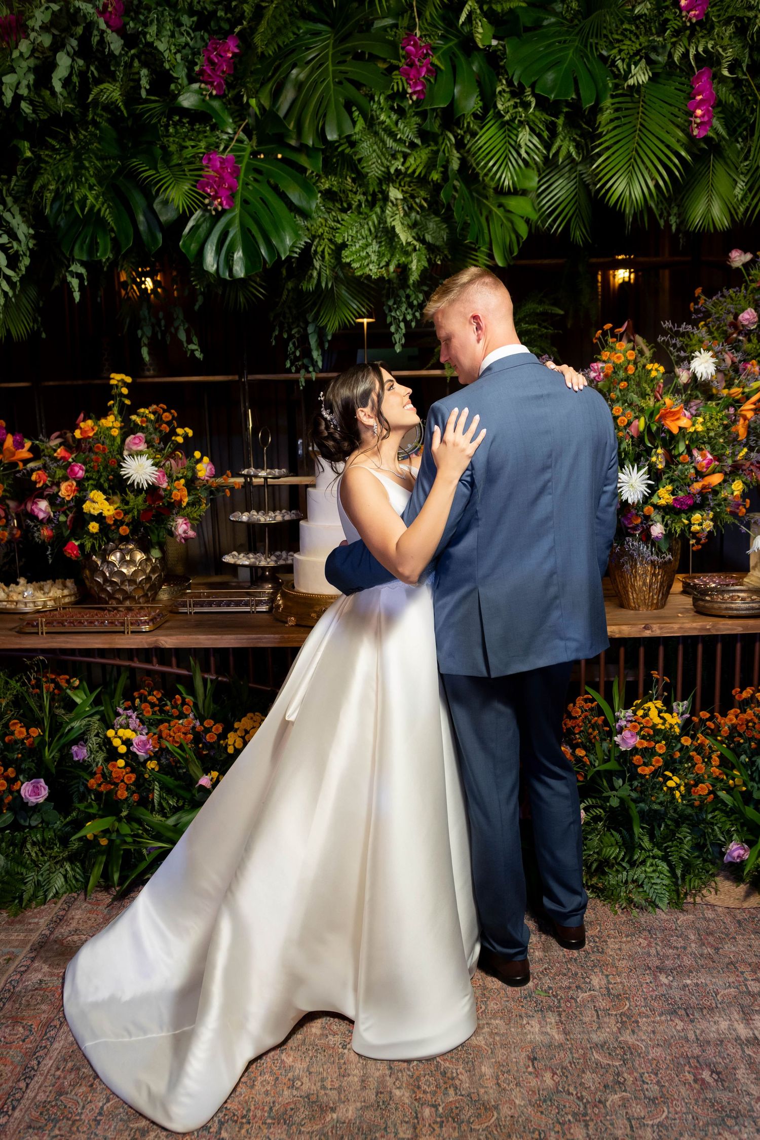 Bride and groom share a romantic dance in front of a lush floral wall with tropical greenery and colorful blooms.